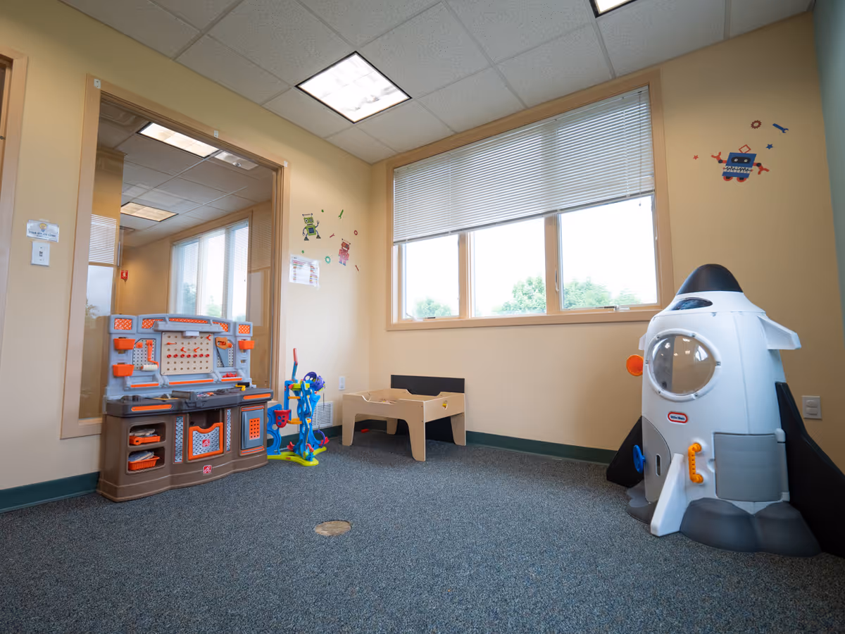 Bright indoor playroom with a plastic kitchen, rocket toy, and window blinds in a facility.