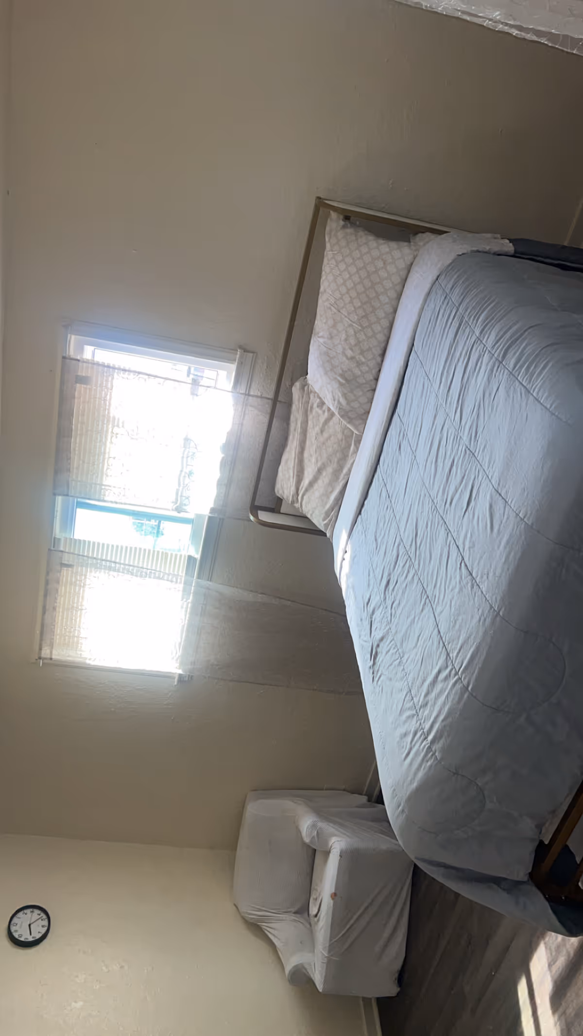 A small bedroom with a neatly made bed, window blinds, and a chair.