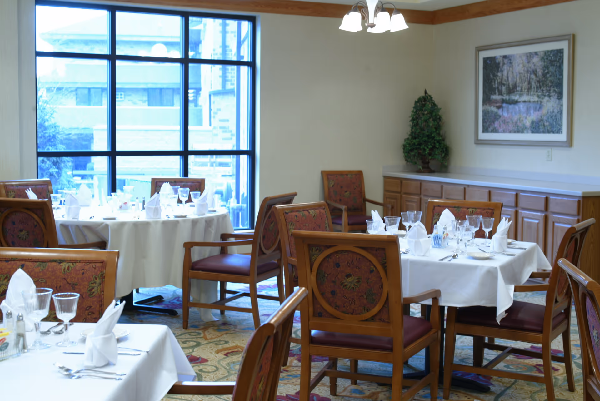 Senior living dining room with round tables set with napkins, glassware, and chairs.