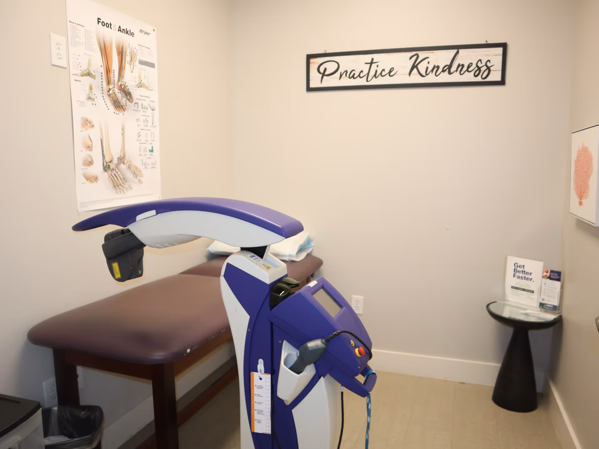 Interior therapy room with a purple medical device, exam table, and 'Practice Kindness' sign.
