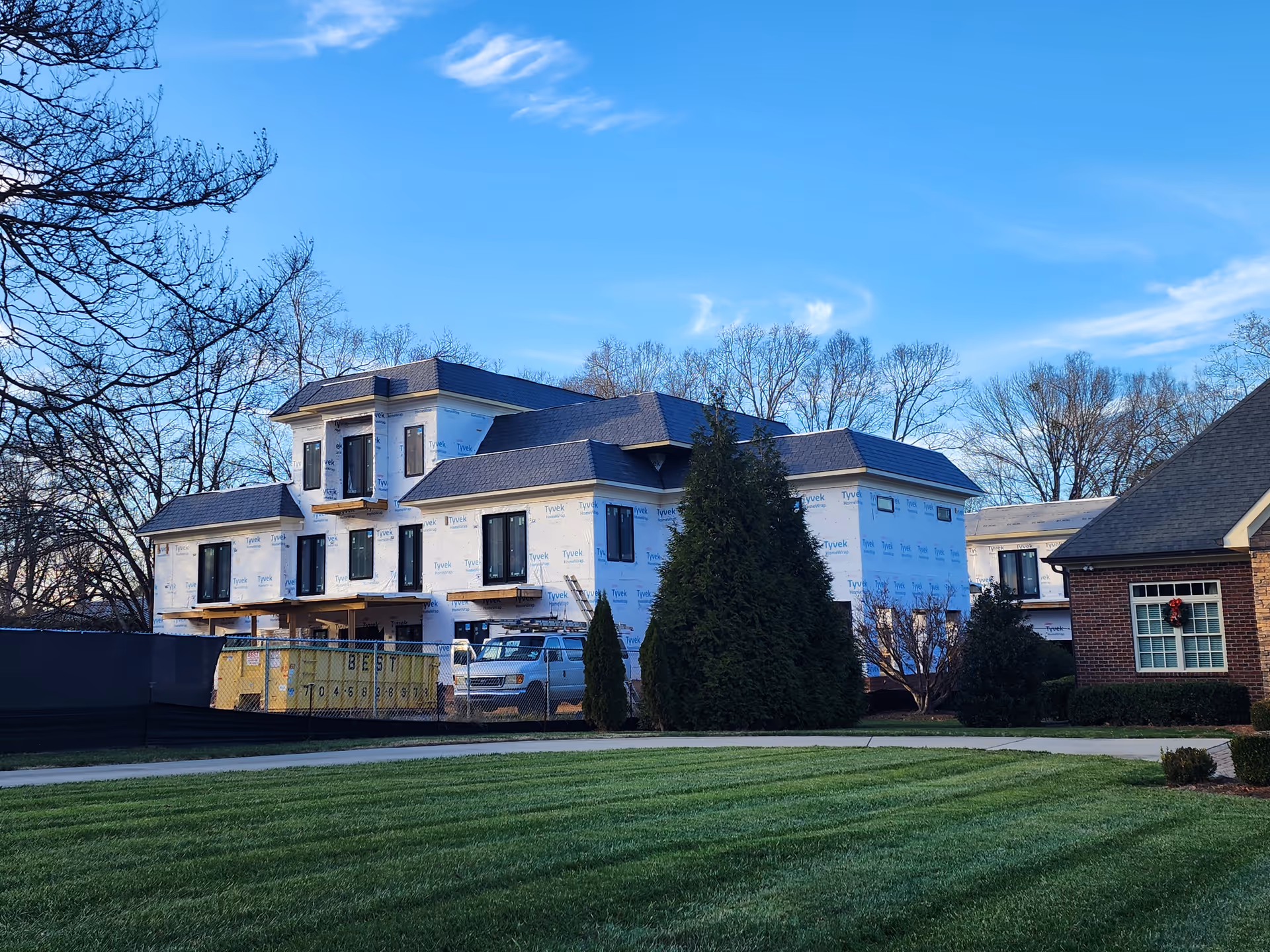 Two-story house under construction wrapped in Tyvek, adjacent to a brick building and green lawn.