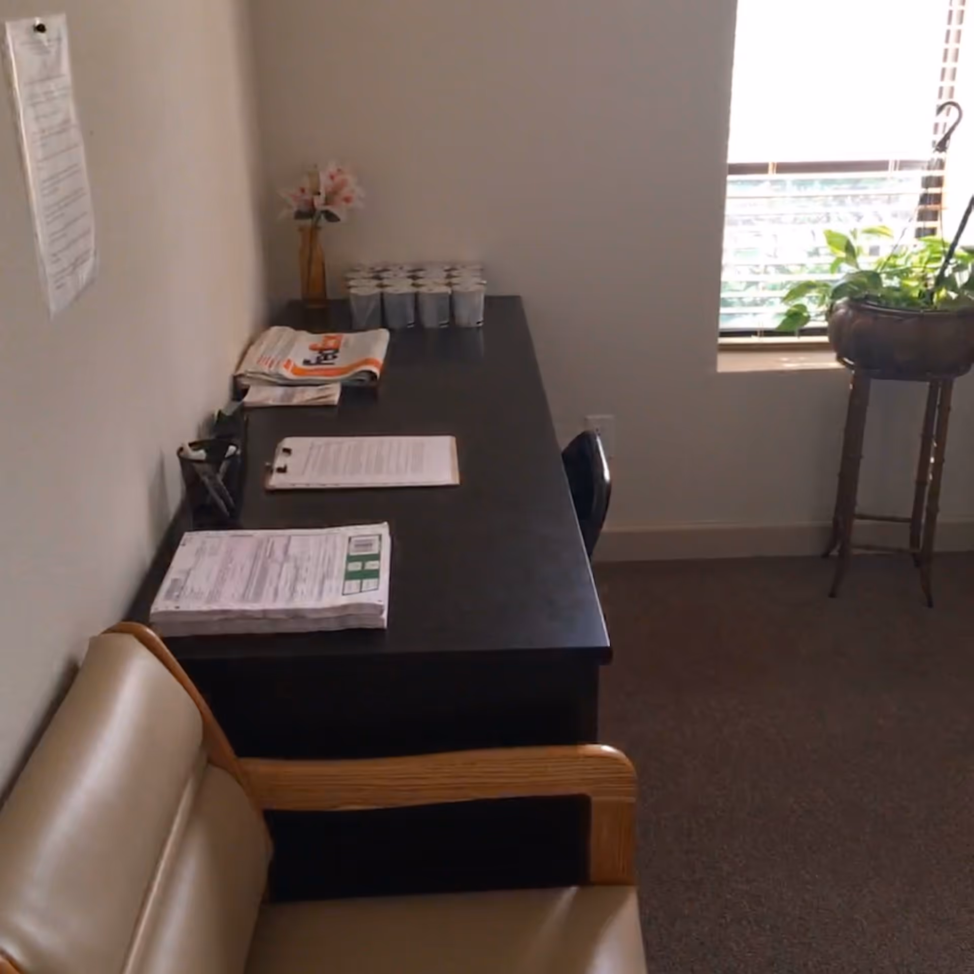A dark desk along a wall with papers, cups, and a plant by a window.