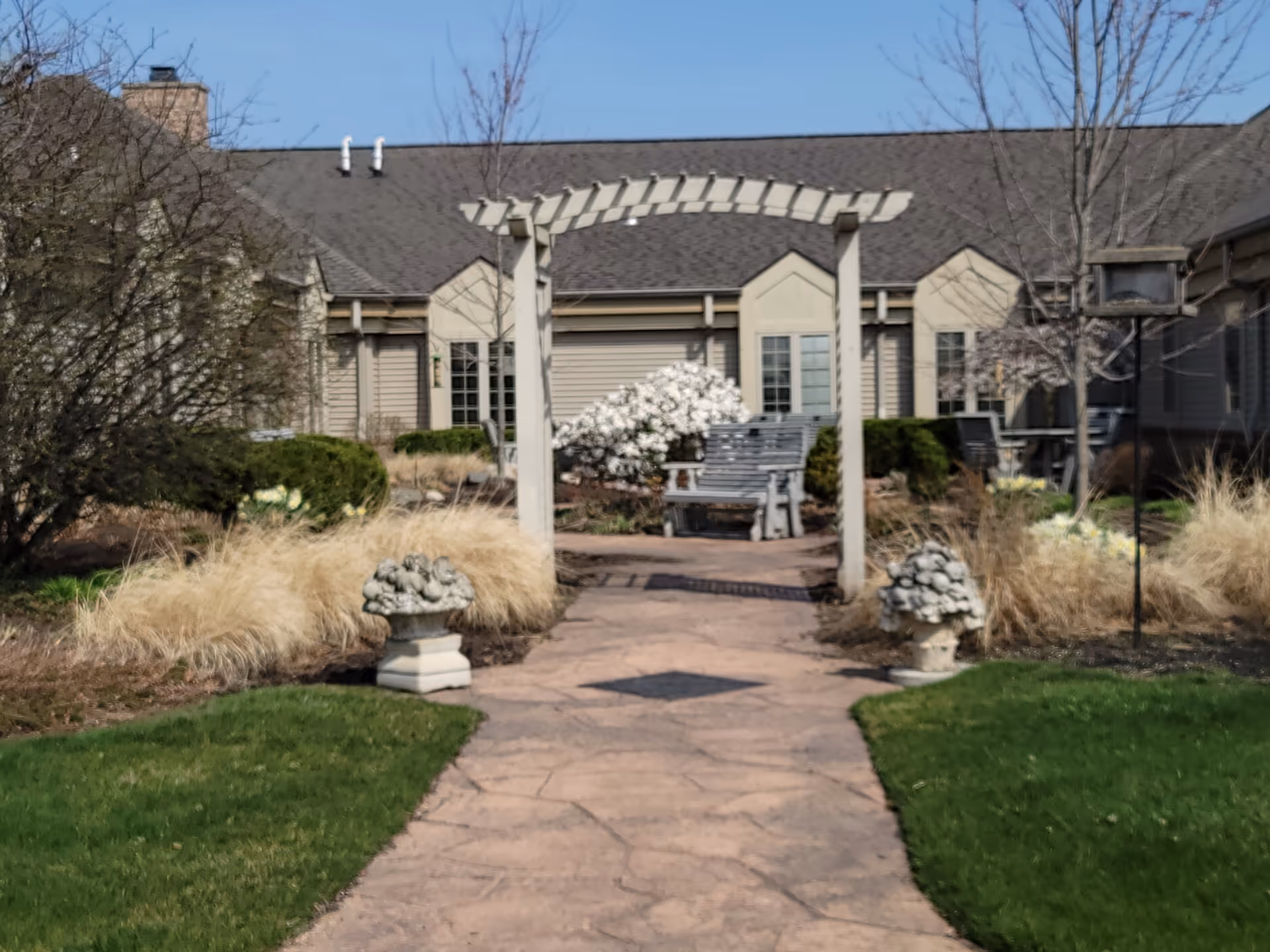 Pathway leads to a courtyard with benches and a pergola in front of senior living facility.