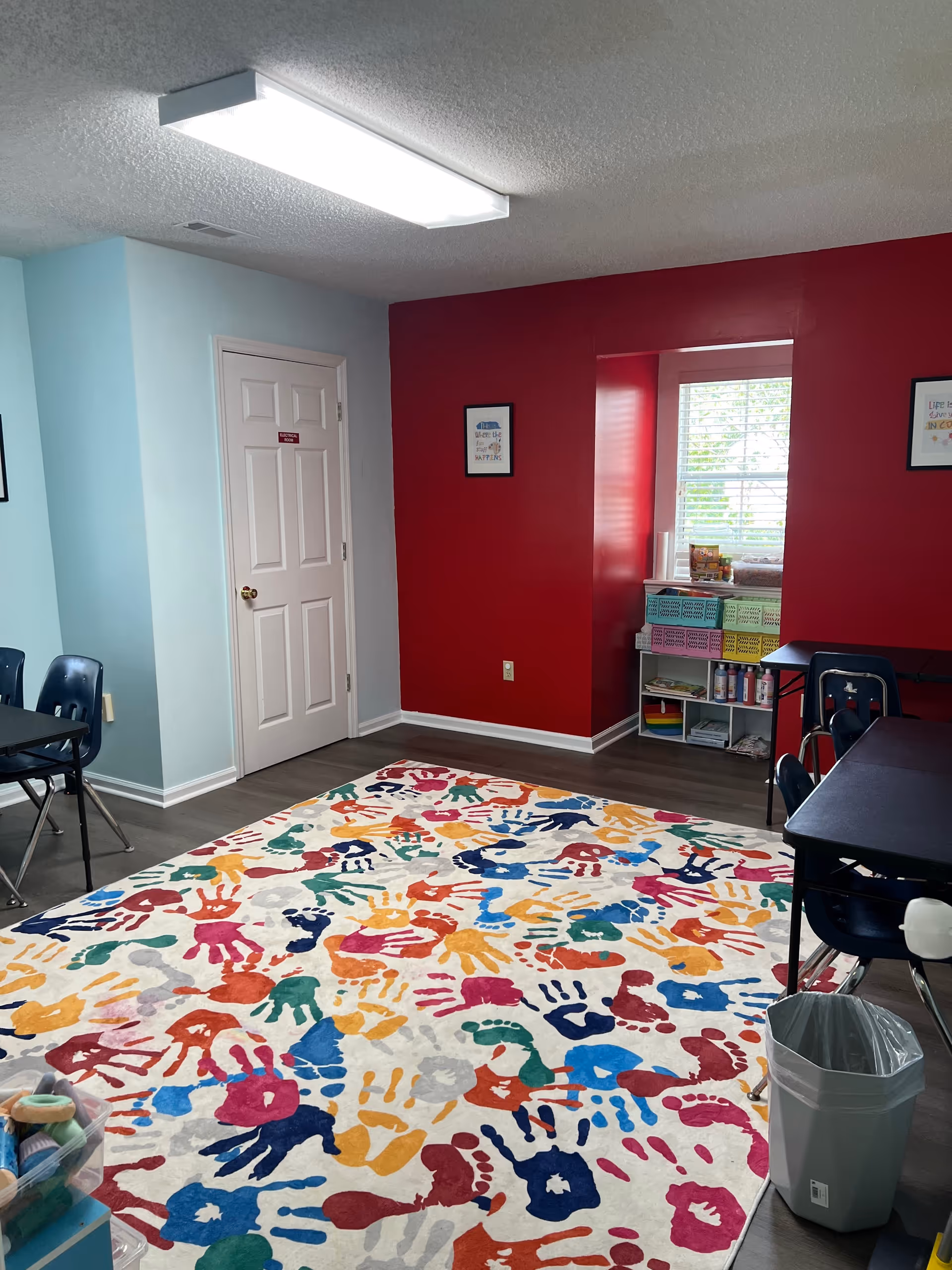 Bright activity room featuring a multicolored handprint rug, tables, chairs, and shelves.