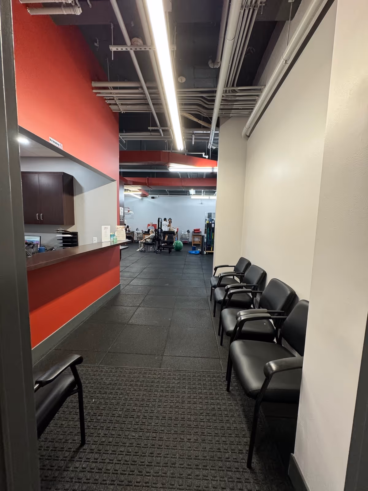 Long clinic corridor with red accent wall, reception desk left, and black chairs along right.