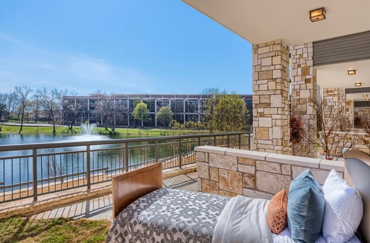 Outdoor balcony with a bed and pillows overlooking a lake, fountain, and walkway.