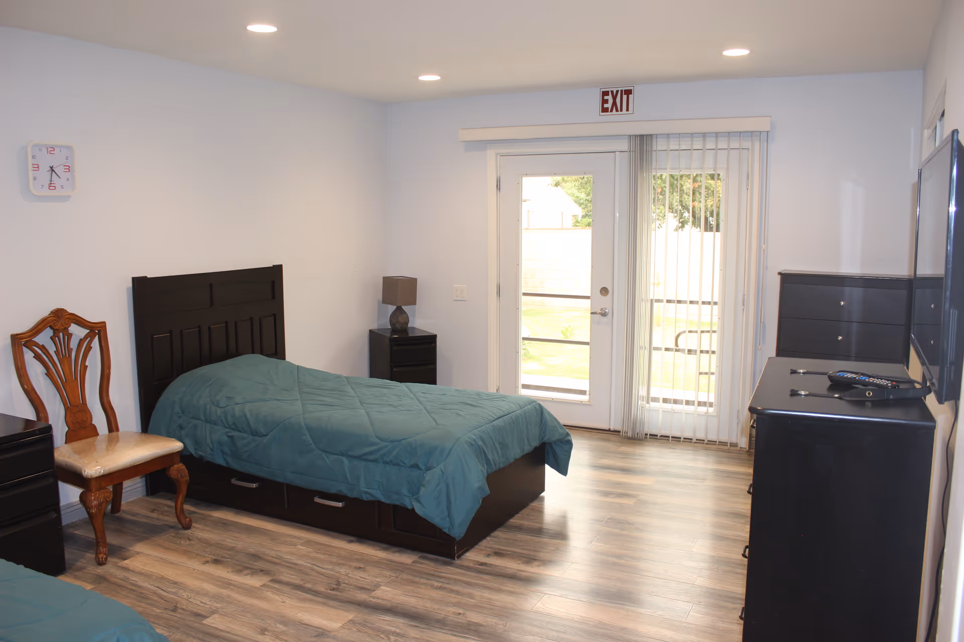 Bedroom in a senior facility with a bed, dresser, chair, and glass door.