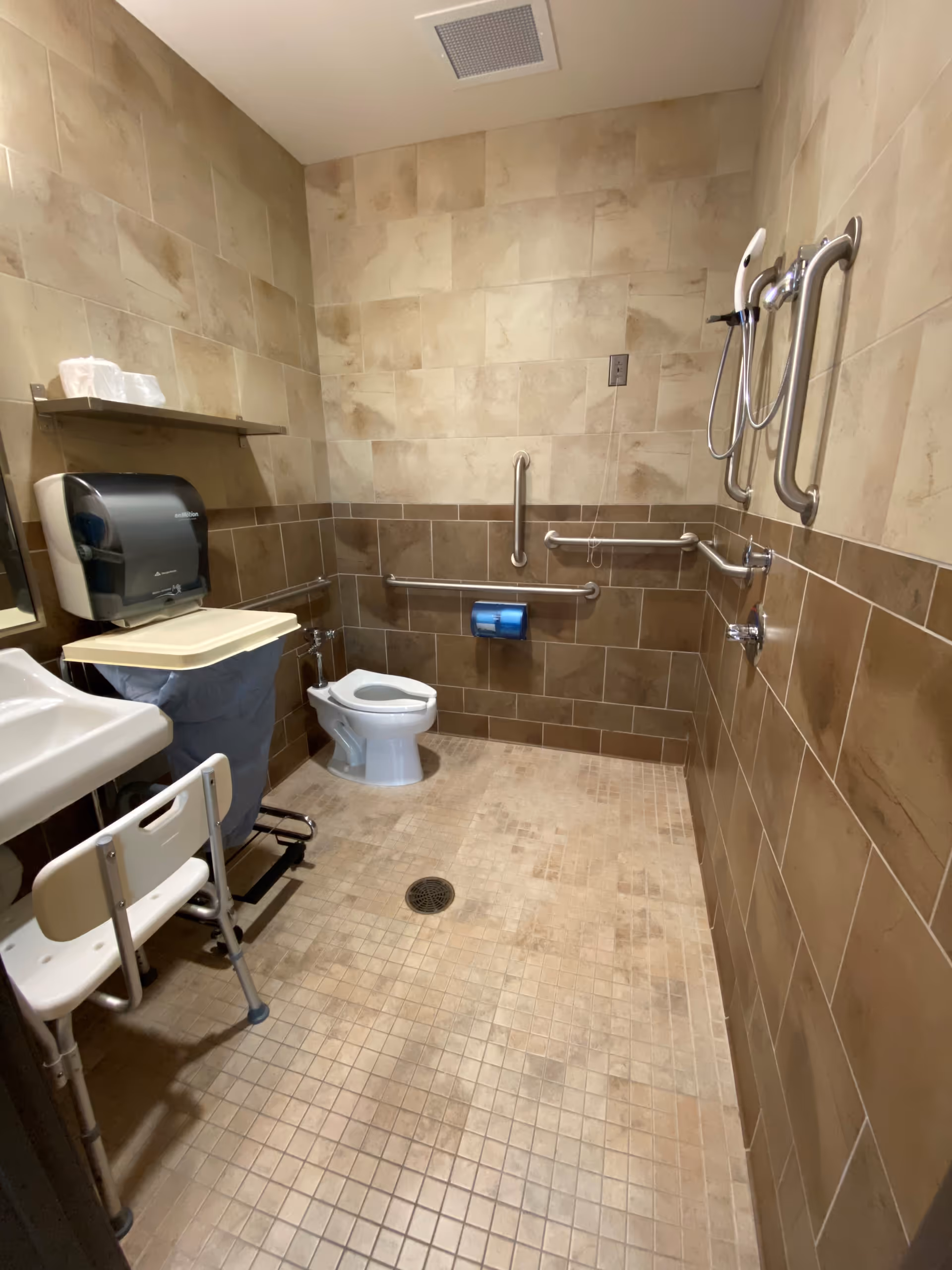 Accessible bathroom featuring grab bars, a toilet, sink, and tiled walls.