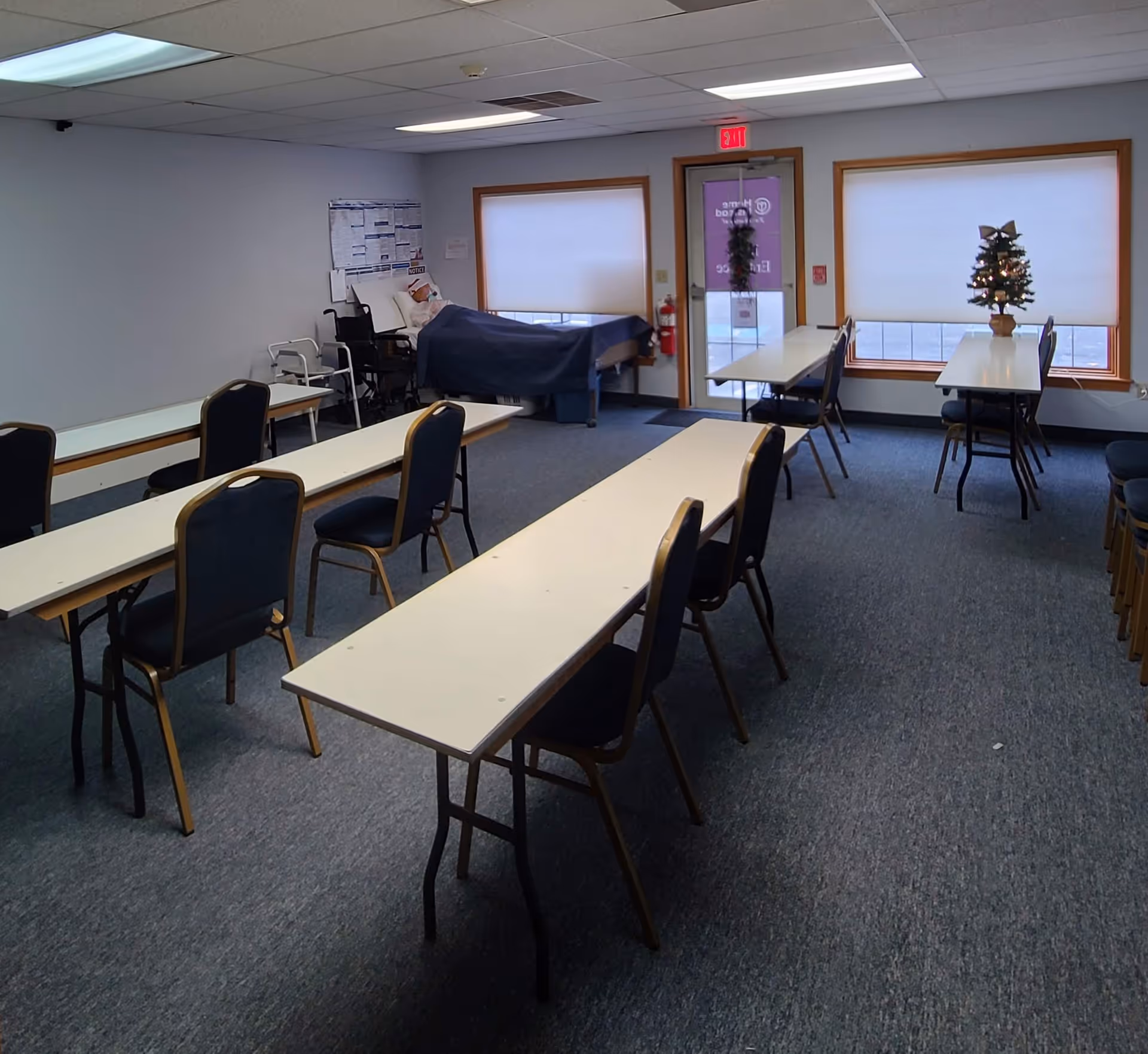 Empty dining room with long tables, chairs, windows, a small Christmas tree, and a hospital bed.