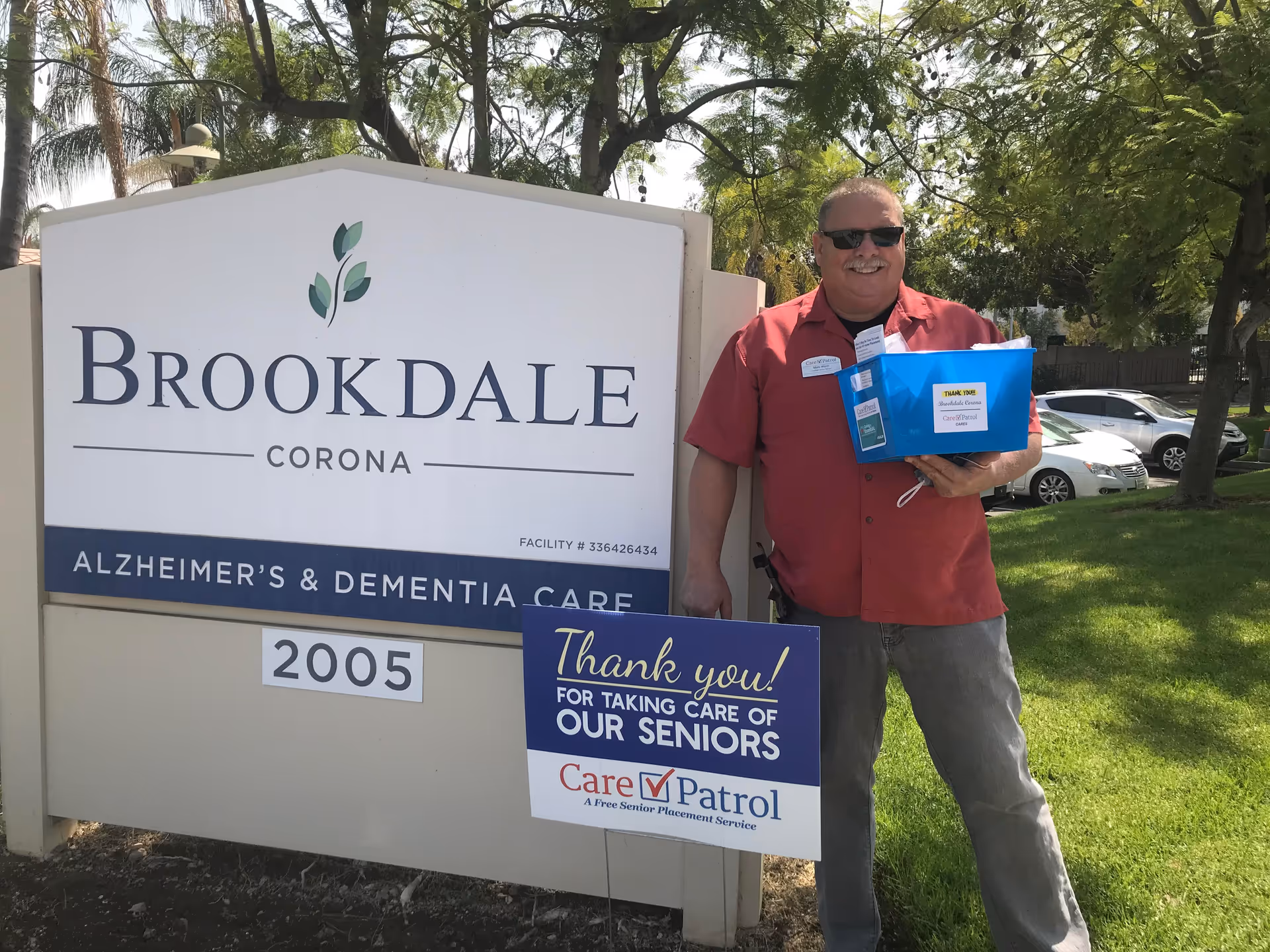 Man in a red shirt stands beside a Brookdale Corona sign outdoors.