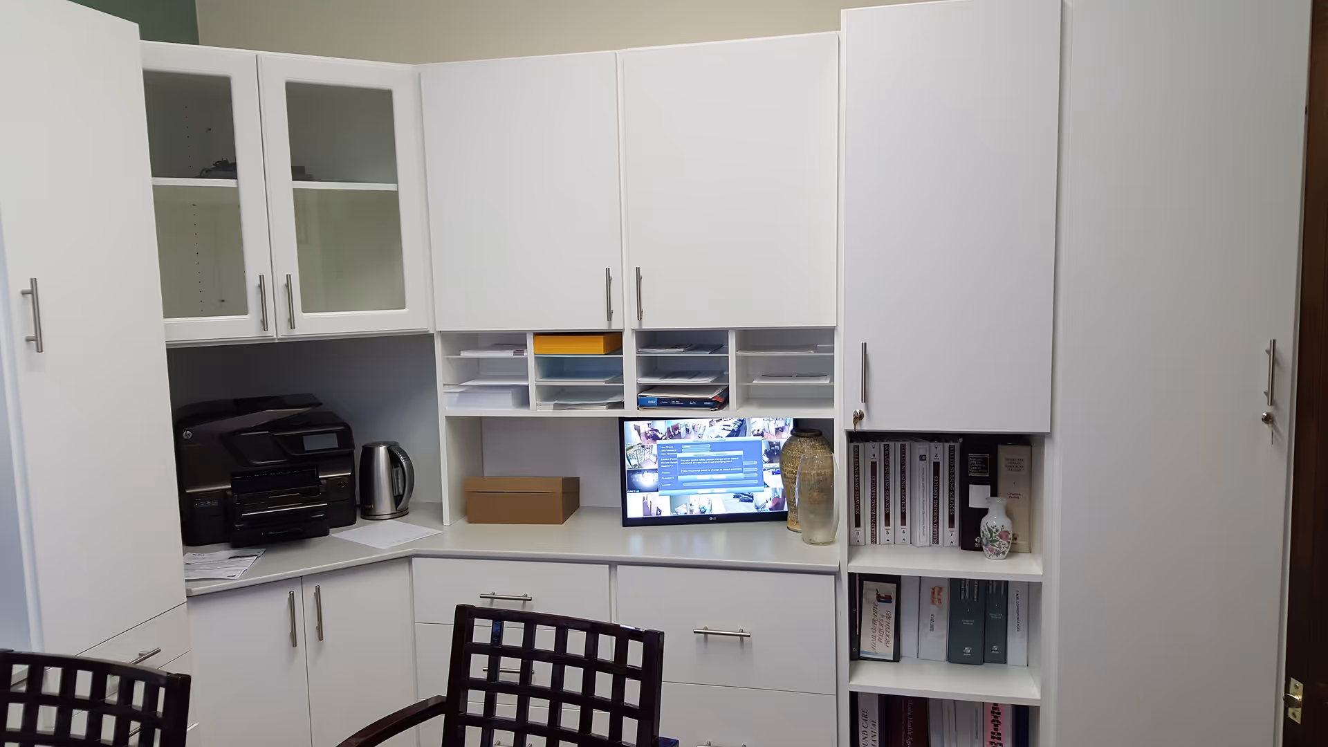 A white-cabinet office area with a desk, computer, printer, kettle, and shelves.
