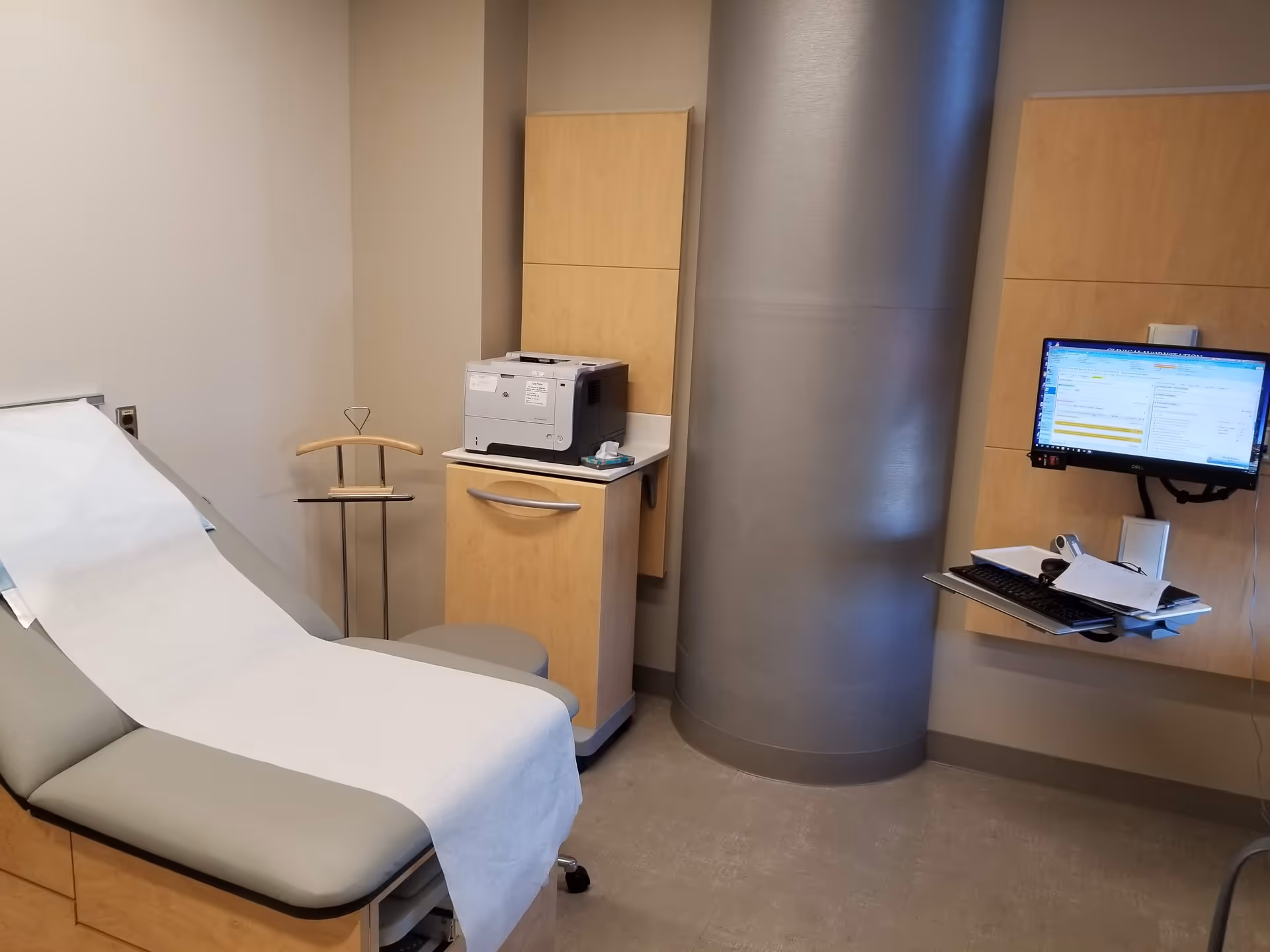 An outpatient exam room with a paper-covered patient chair, printer, cabinets, and a wall-mounted computer.