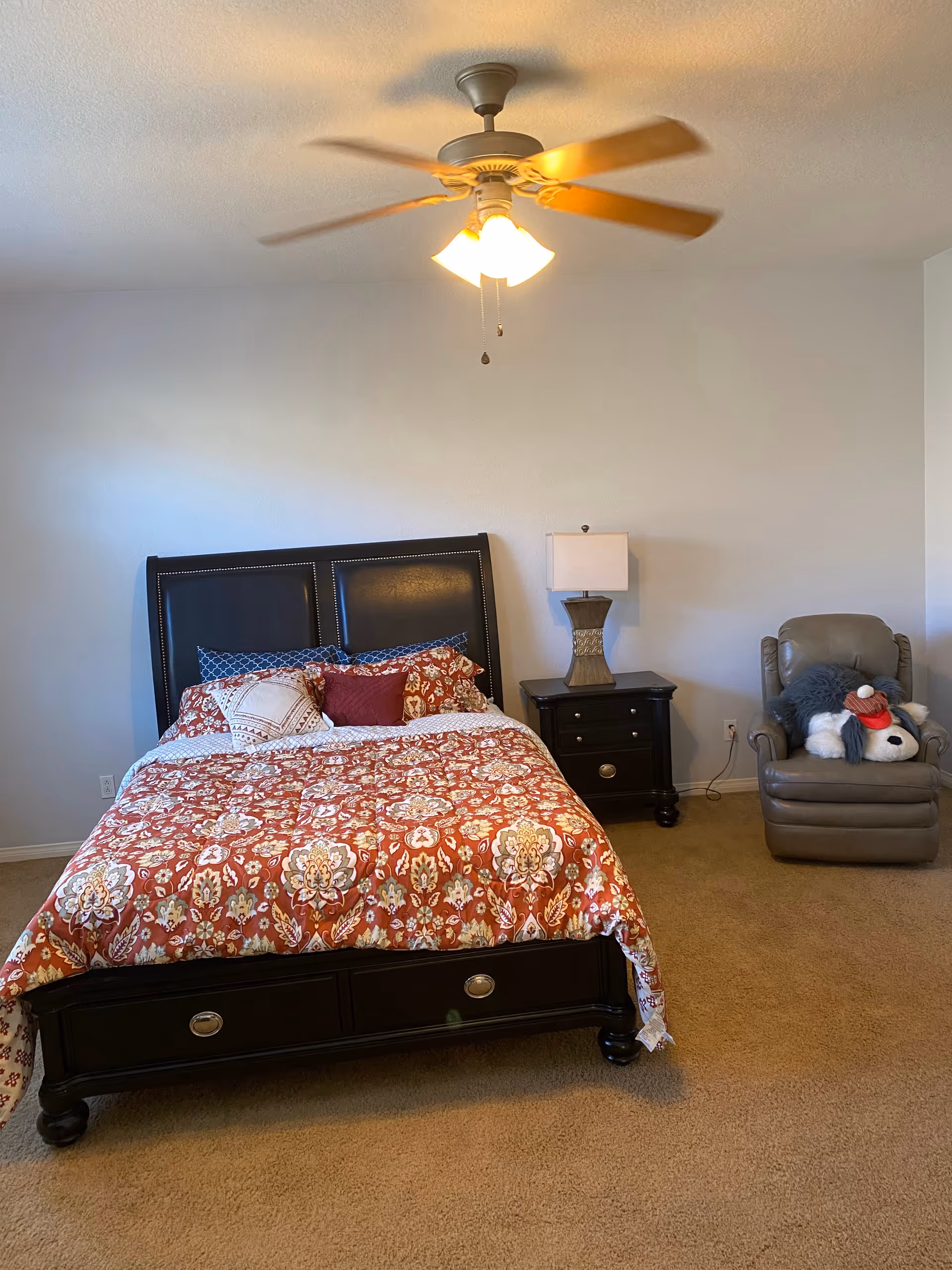 A furnished bedroom with a dark bed, nightstand, ceiling fan, and an armchair.