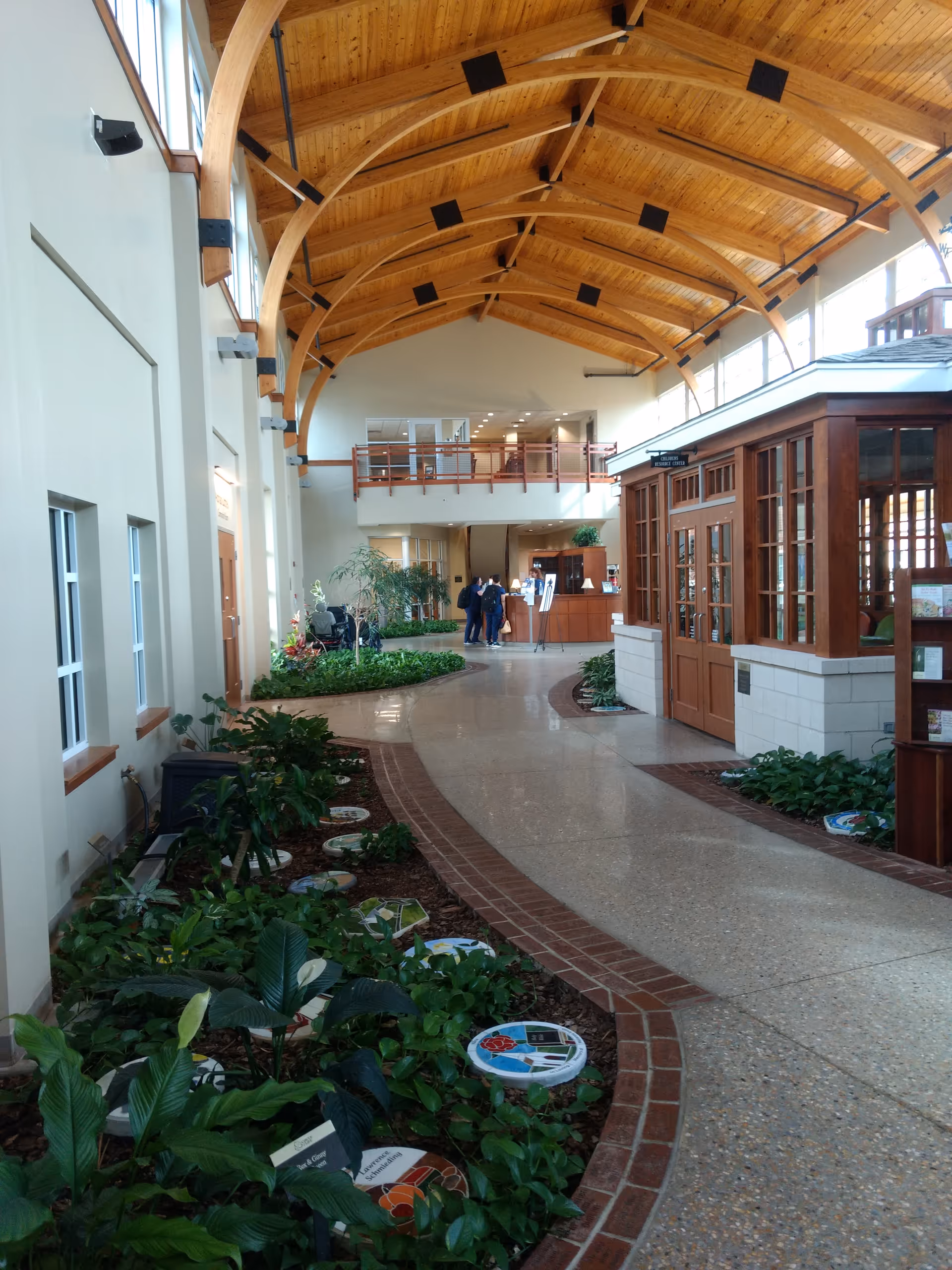 Spacious indoor atrium with wooden vaulted ceiling, plants lining a curved walkway, and a reception desk.
