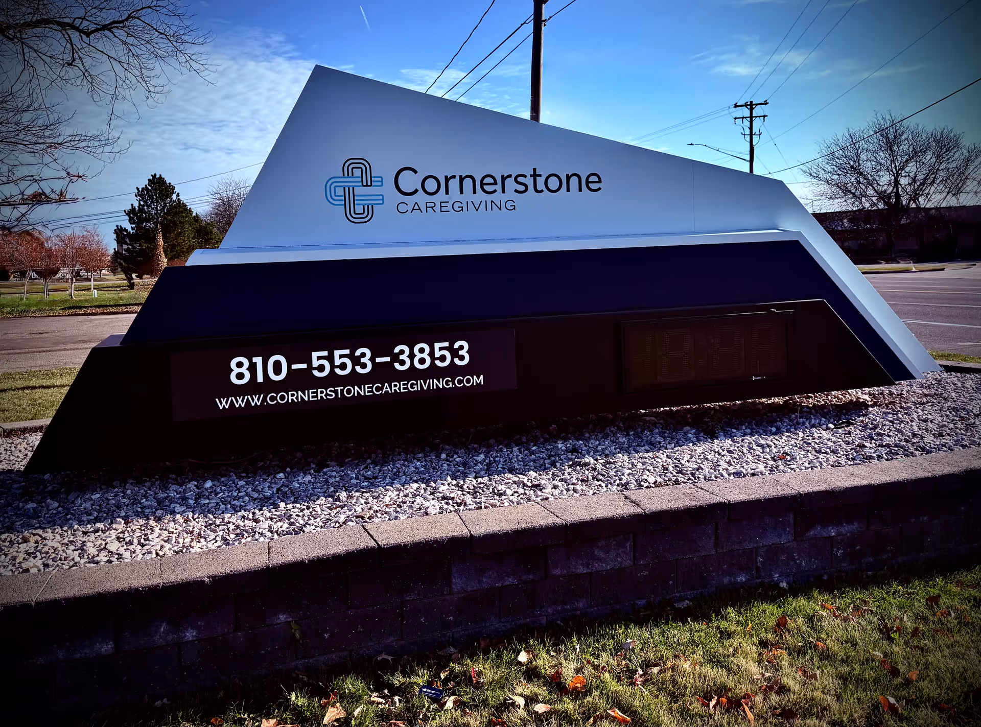 An exterior Cornerstone Caregiving sign sits on a rock-filled, landscaped bed by the road.