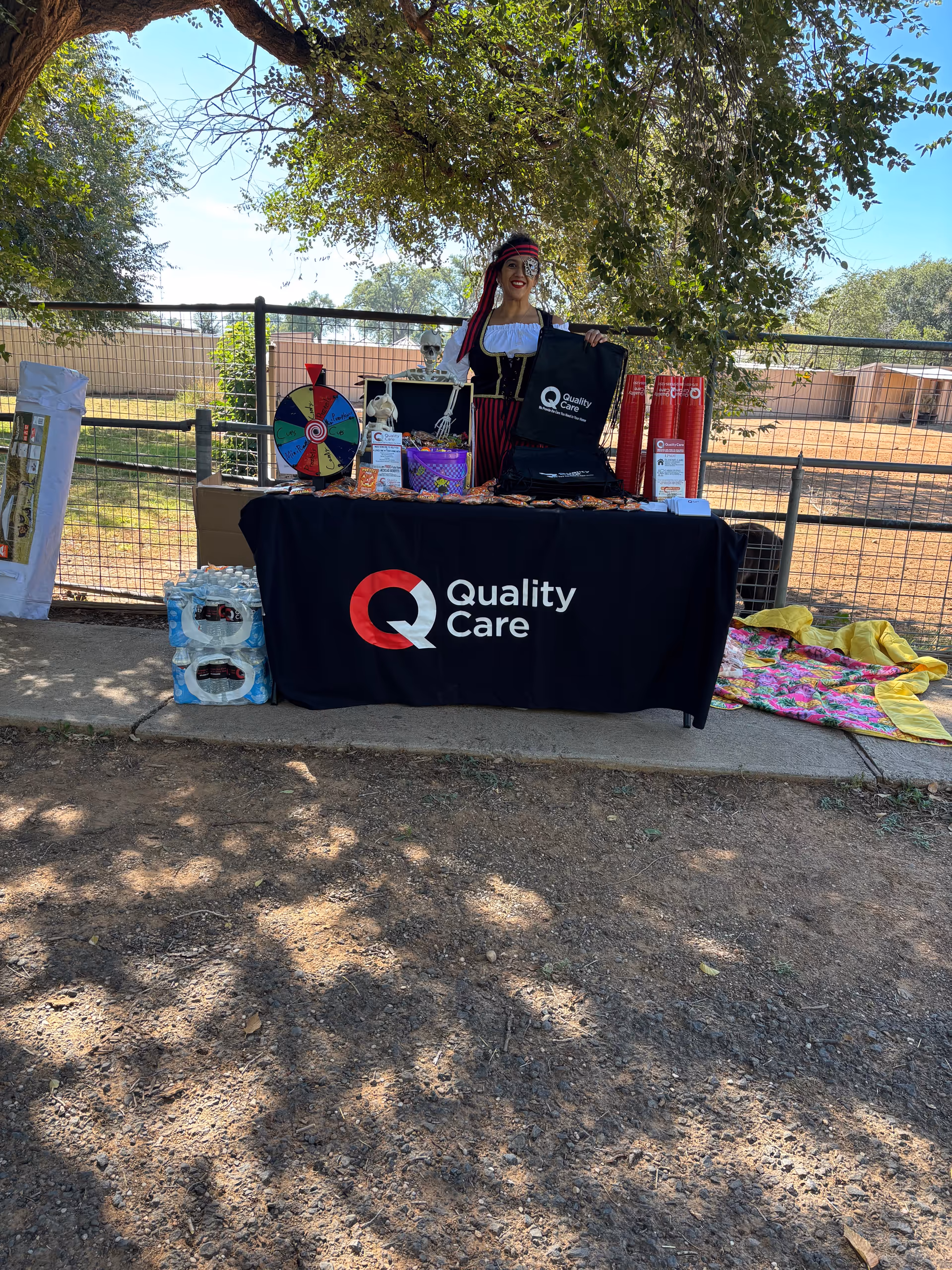 Outdoor table with Quality Care branding and a costumed presenter under a shaded tree.
