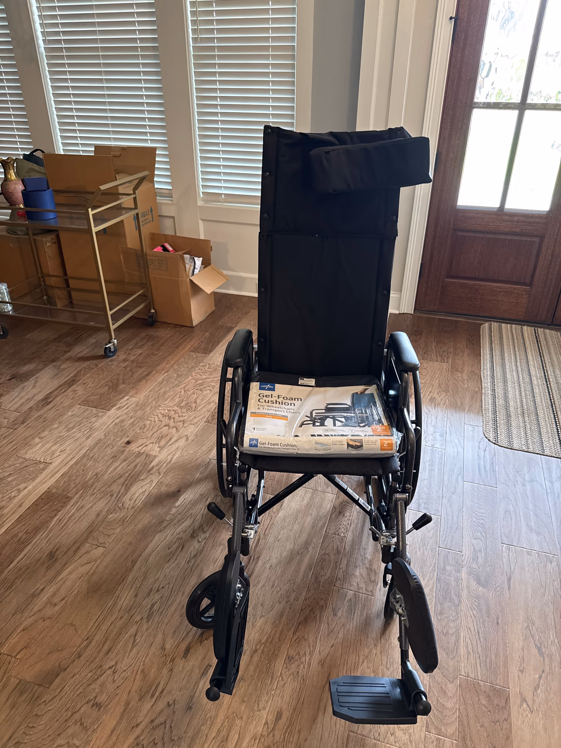 A wheelchair sits inside a room with boxes and a rolling cart nearby.