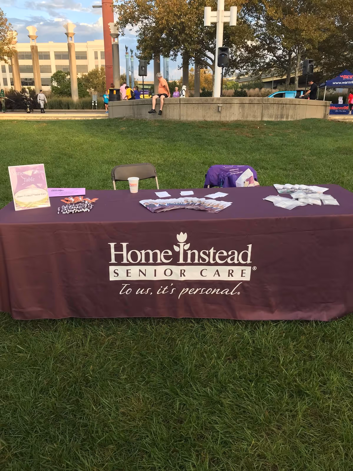 A purple Home Instead table on a grassy outdoor area with people in background.