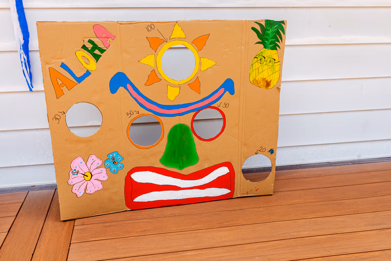 Colorful cardboard face cutout with sun, pineapple, and flowers on an outdoor deck.