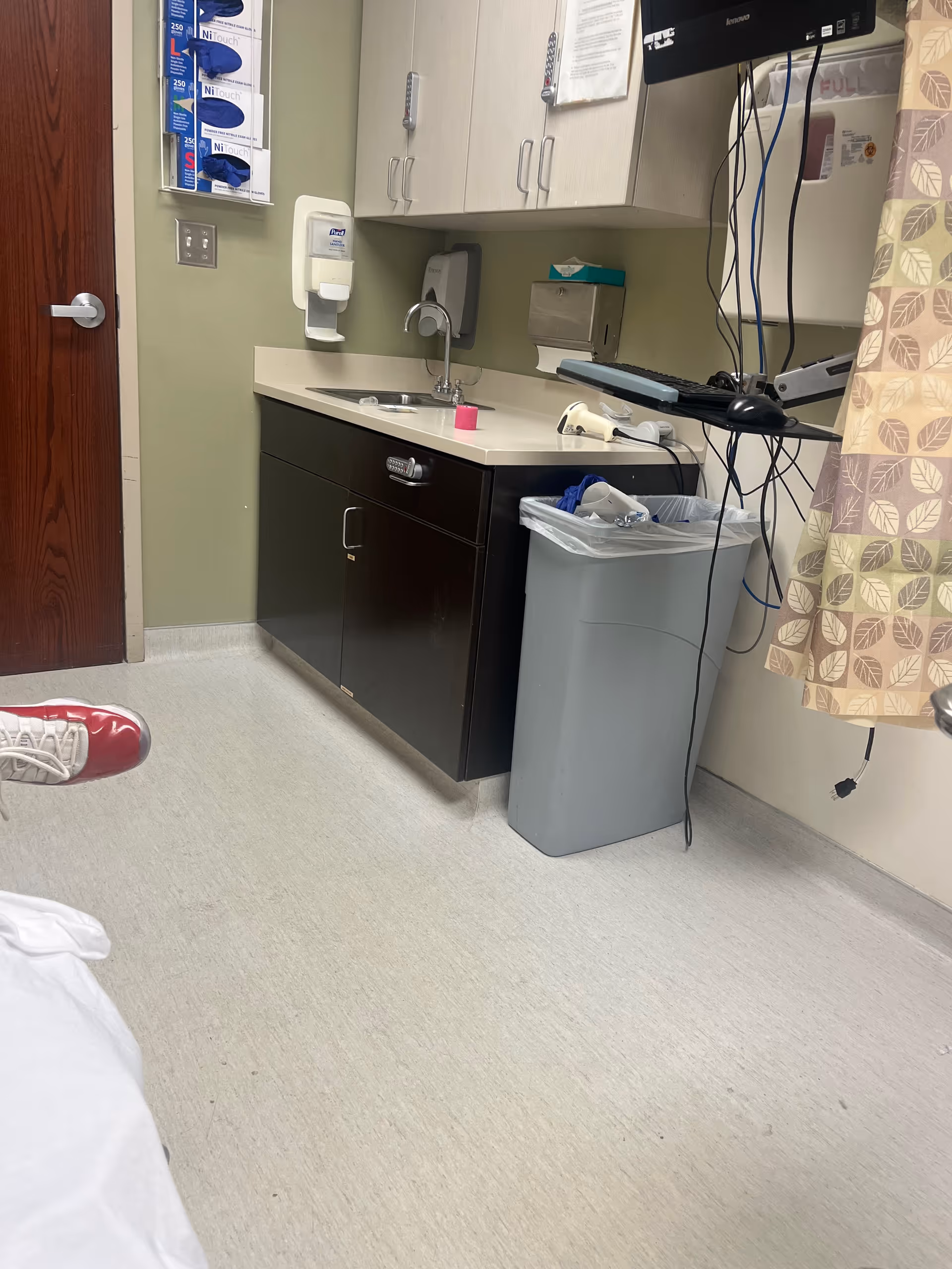Small hospital kitchenette area with sink, cabinets, trash can, and computer monitor.