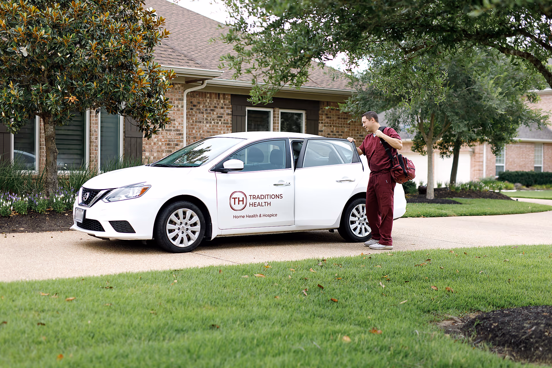 A caregiver in maroon scrubs approaches a white car with a medical logo outside brick facility.