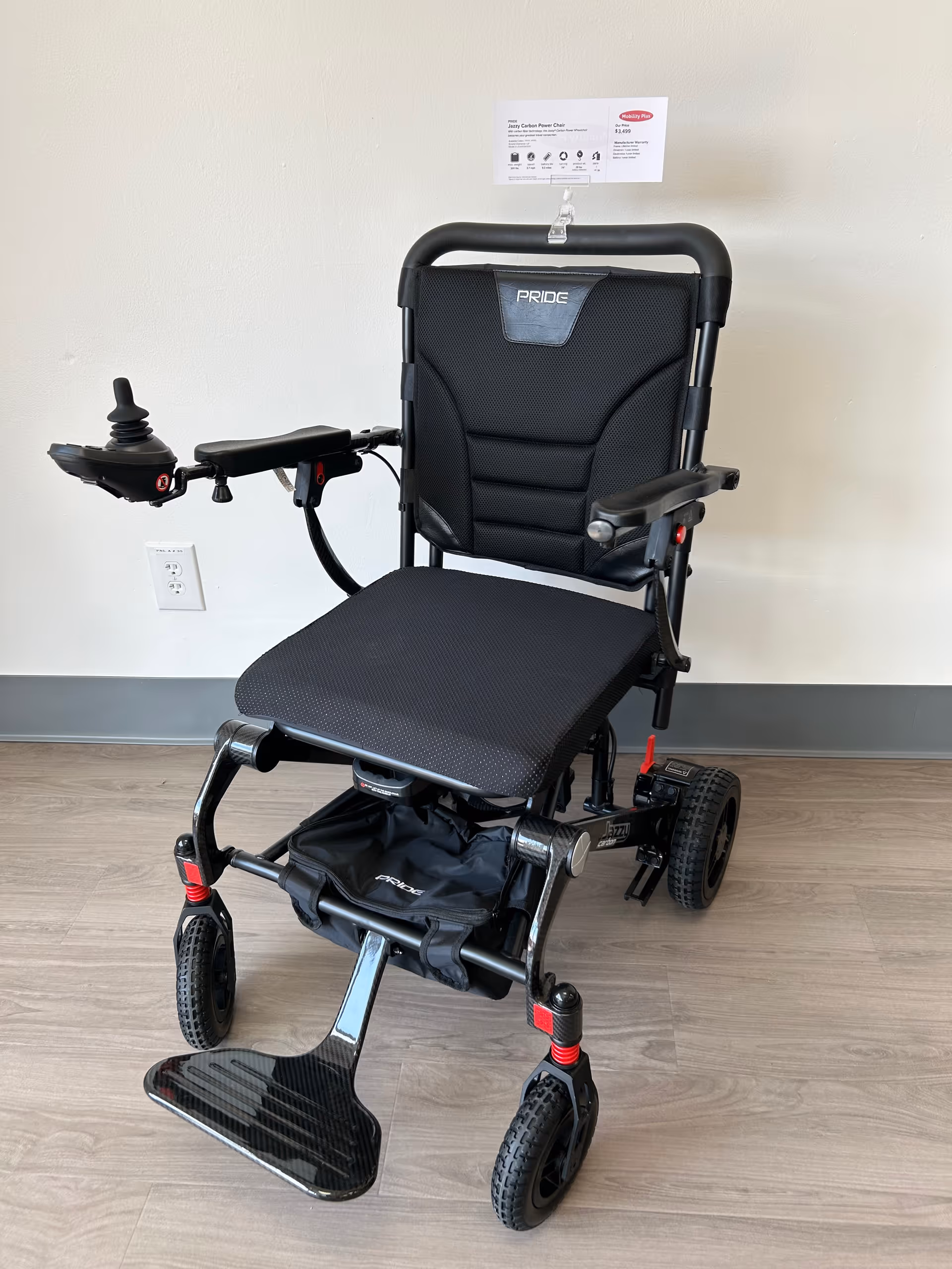 Black mobility wheelchair with joystick, parked in a clean indoor hallway of a senior living facility.