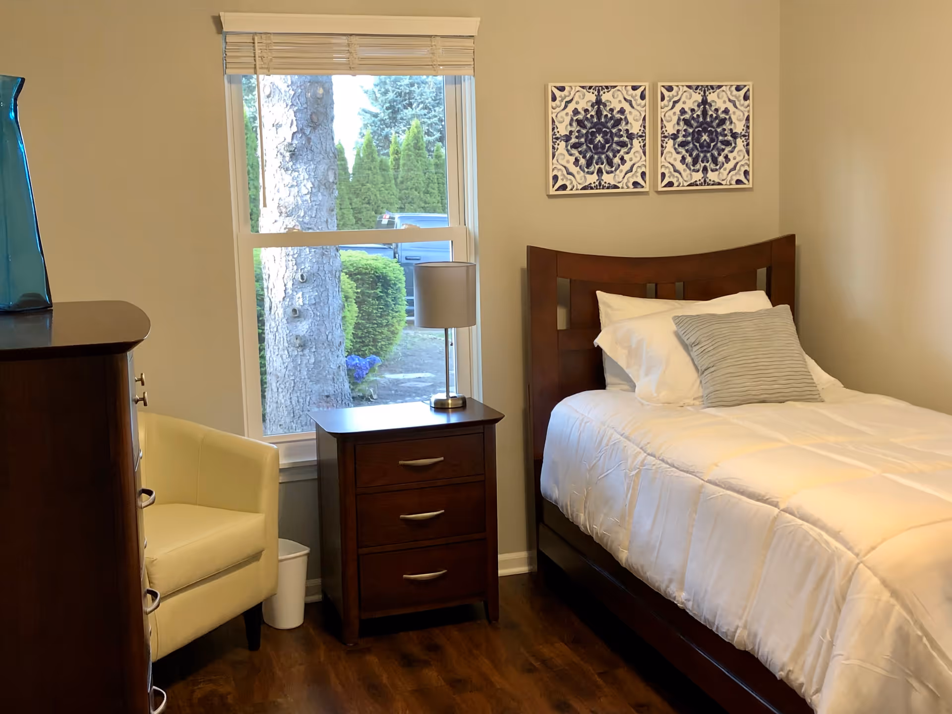 A tidy bedroom in a senior facility with a wooden bed, dresser, chair, and window.