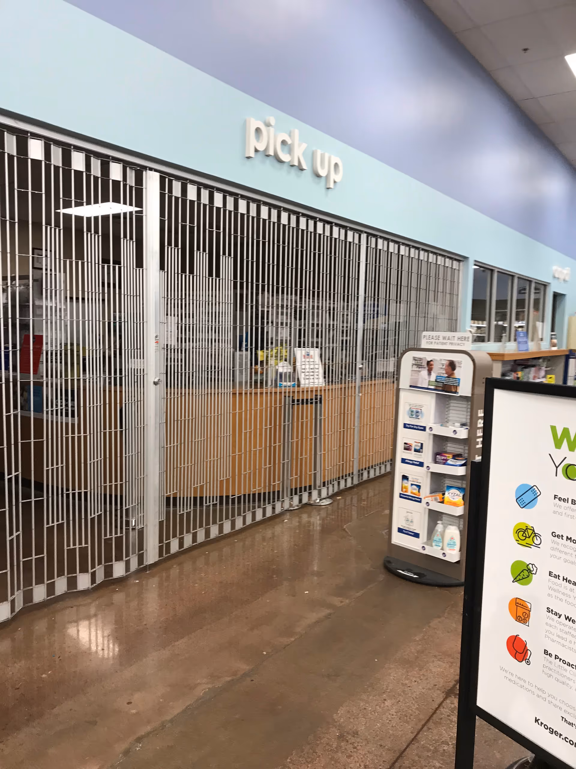 Inside Kroger Pharmacy pickup area with metal security gates, counter behind glass, signage and displays.