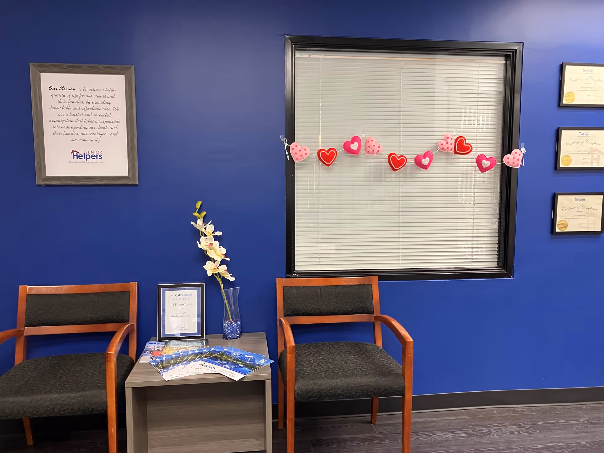 Blue-walled waiting area with two wooden chairs, a small table, a window, and heart garland.