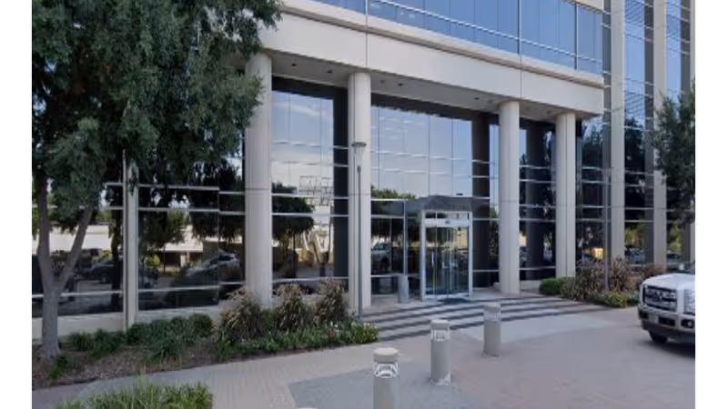 Exterior view of a modern glass-front building entrance with columns and landscaping.