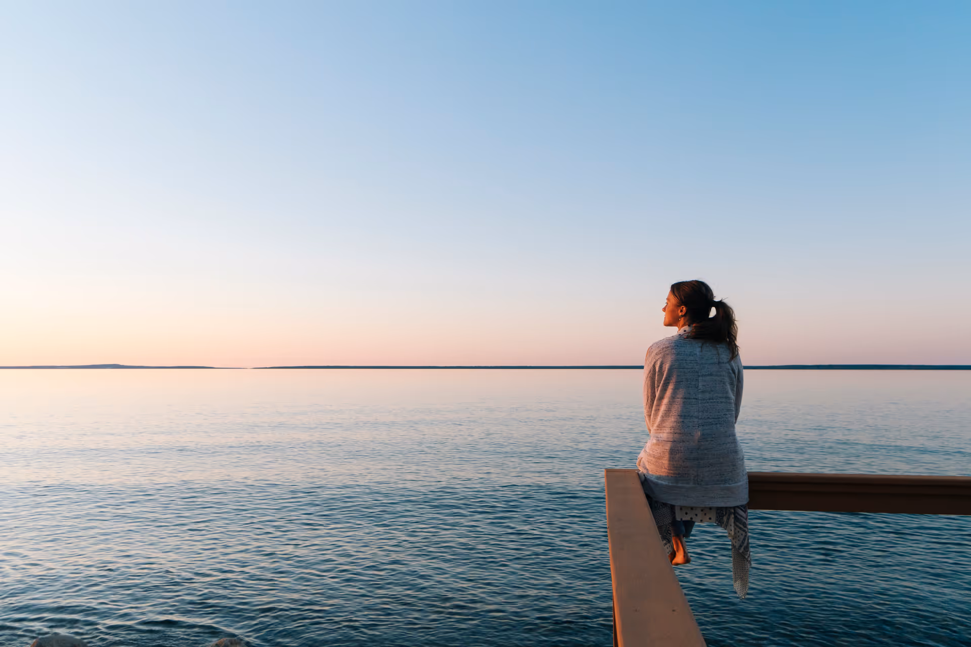 A woman sits on a railing overlooking calm water at sunset.