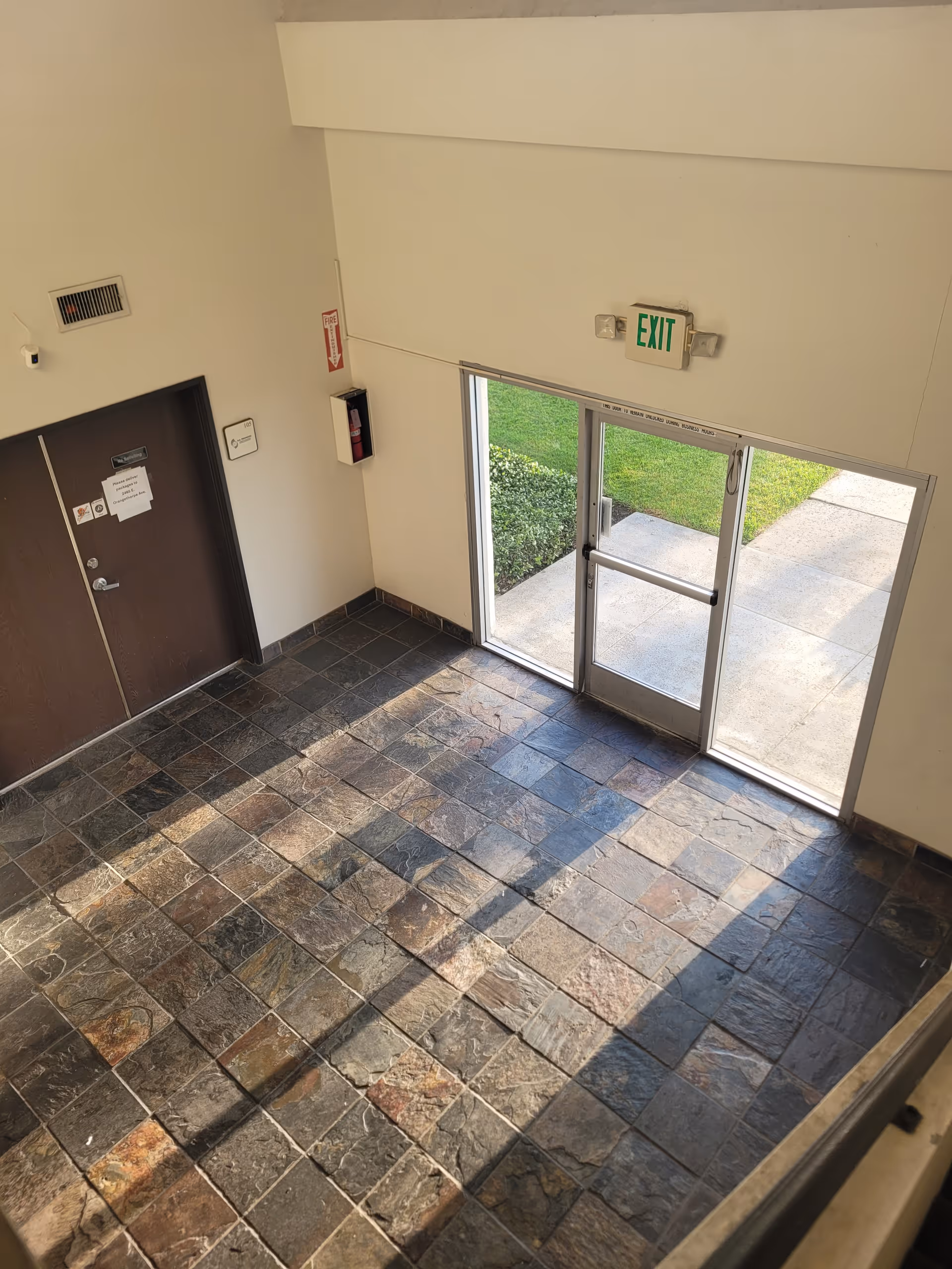 A tiled lobby with glass exit doors, an exit sign, and outdoor greenery beyond.