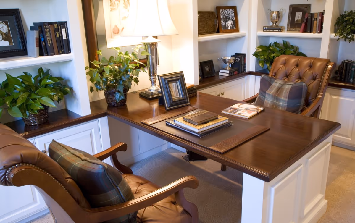Polished wooden executive desk with leather chairs, bookshelves, lamps, and plants in an office.