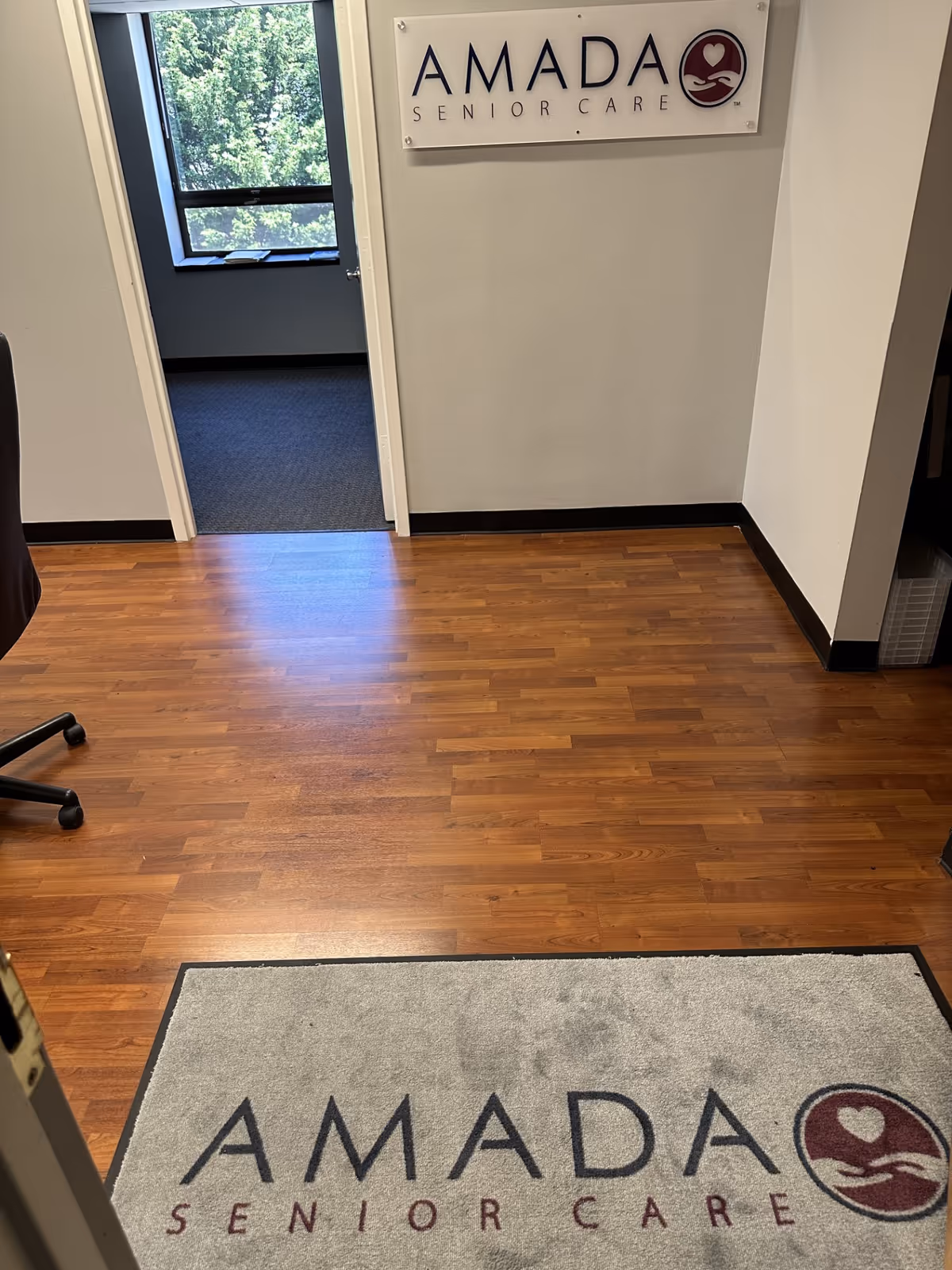 Interior hallway of Amada Senior Care with sign, wood floor, doorway to an office.