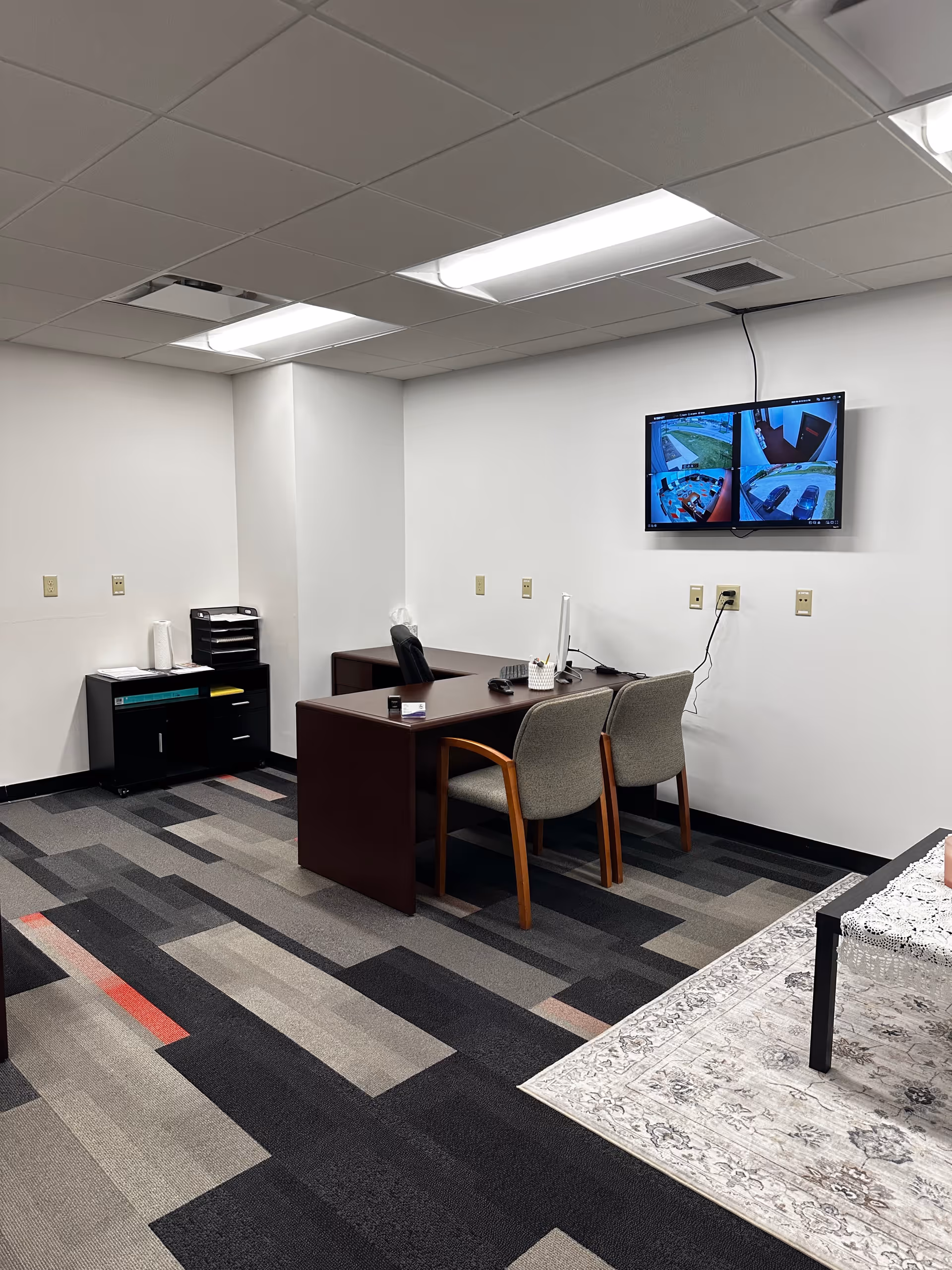 Office desk with two gray chairs and a wall-mounted monitor displaying security feeds.