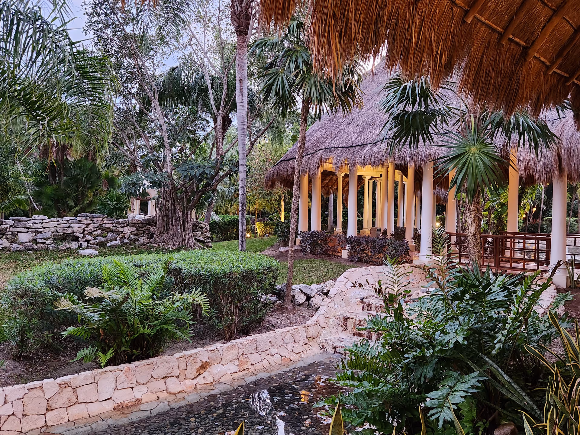Outdoor tropical garden with a thatched-roof pavilion, stone pathways, and lush greenery.