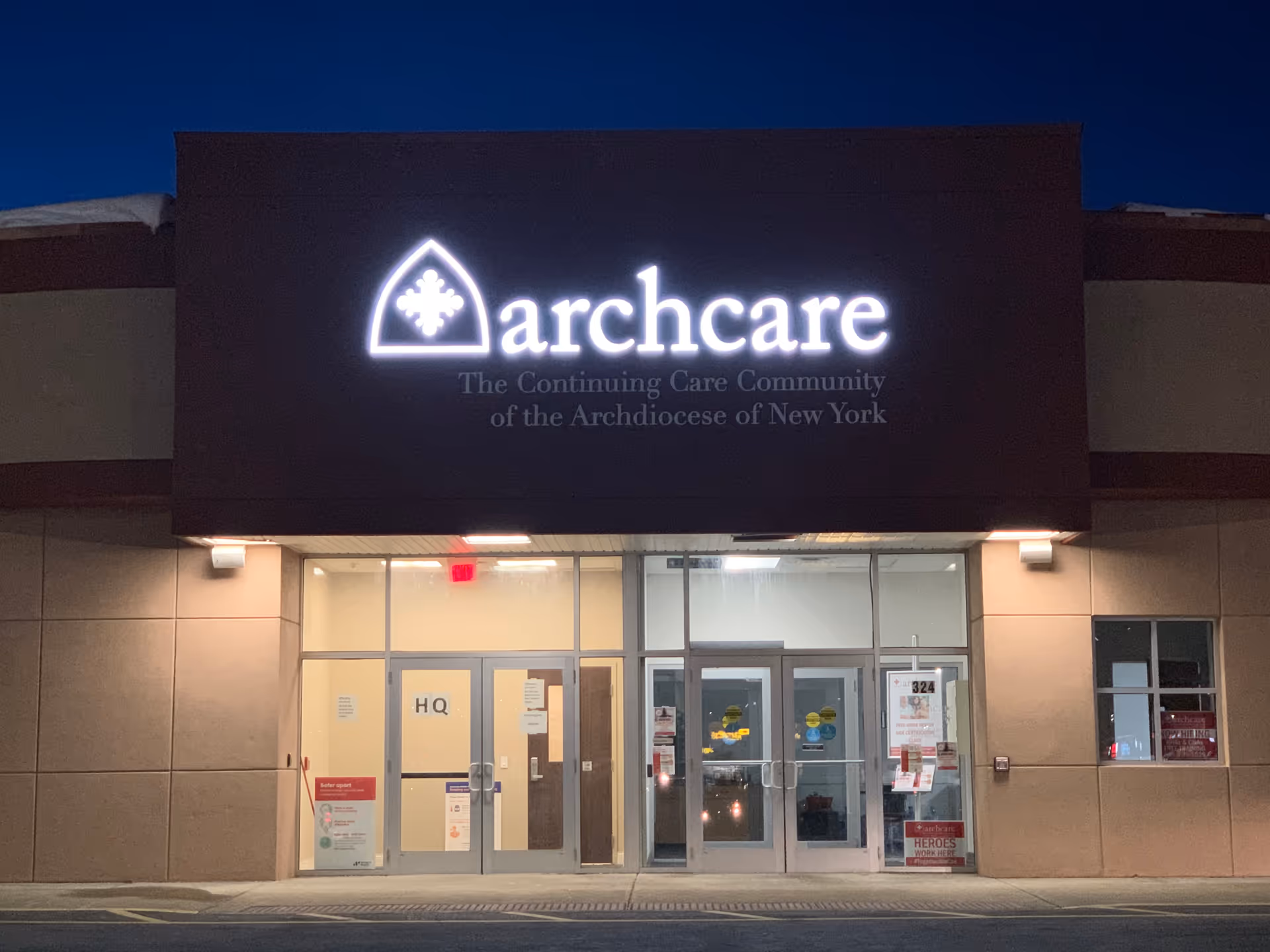 Nighttime exterior of a building with a lit ArchCare sign above glass entry doors.