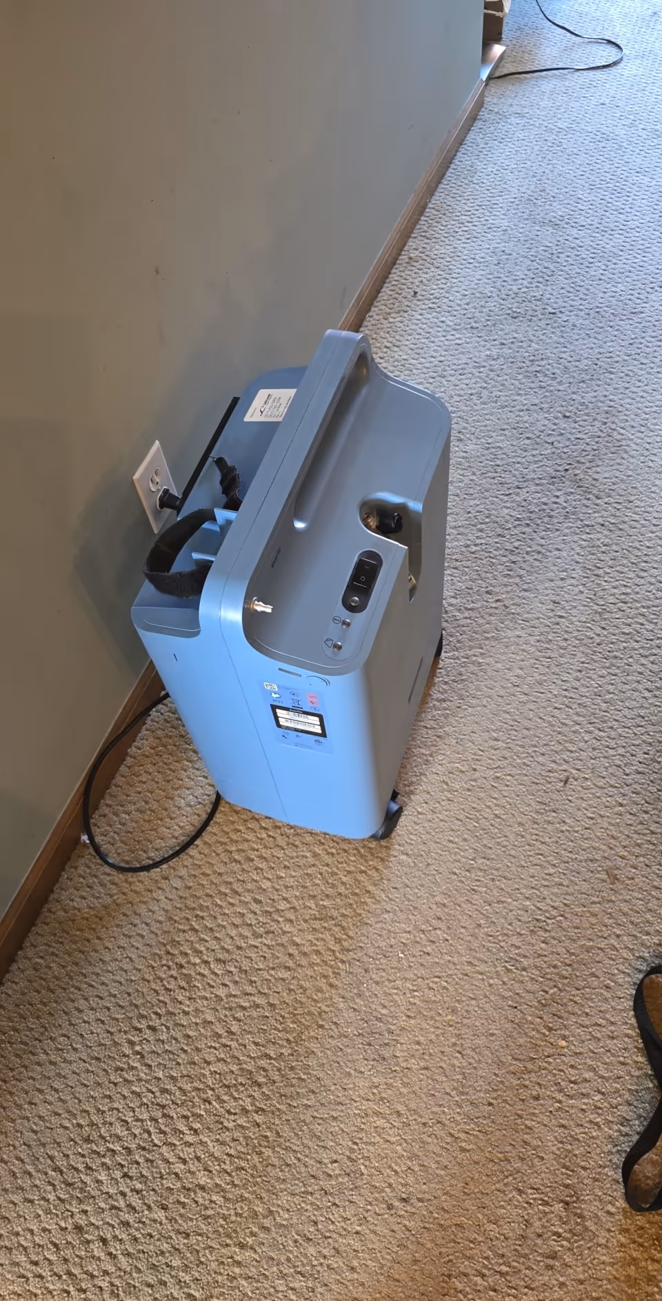 Blue oxygen concentrator in a carpeted hallway near a wall outlet.
