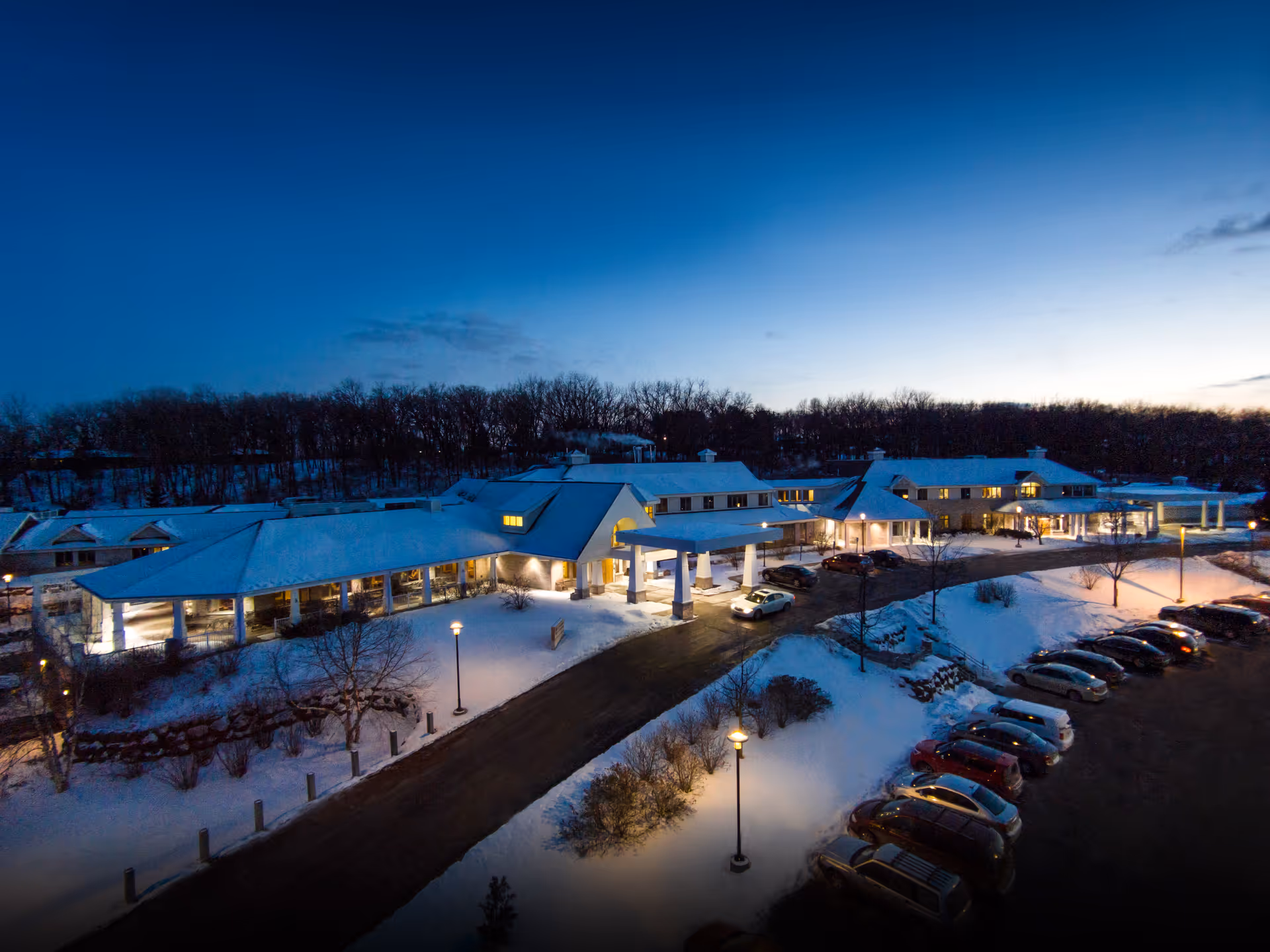 A snow-covered senior living facility exterior at dusk with a parking lot and entrance.