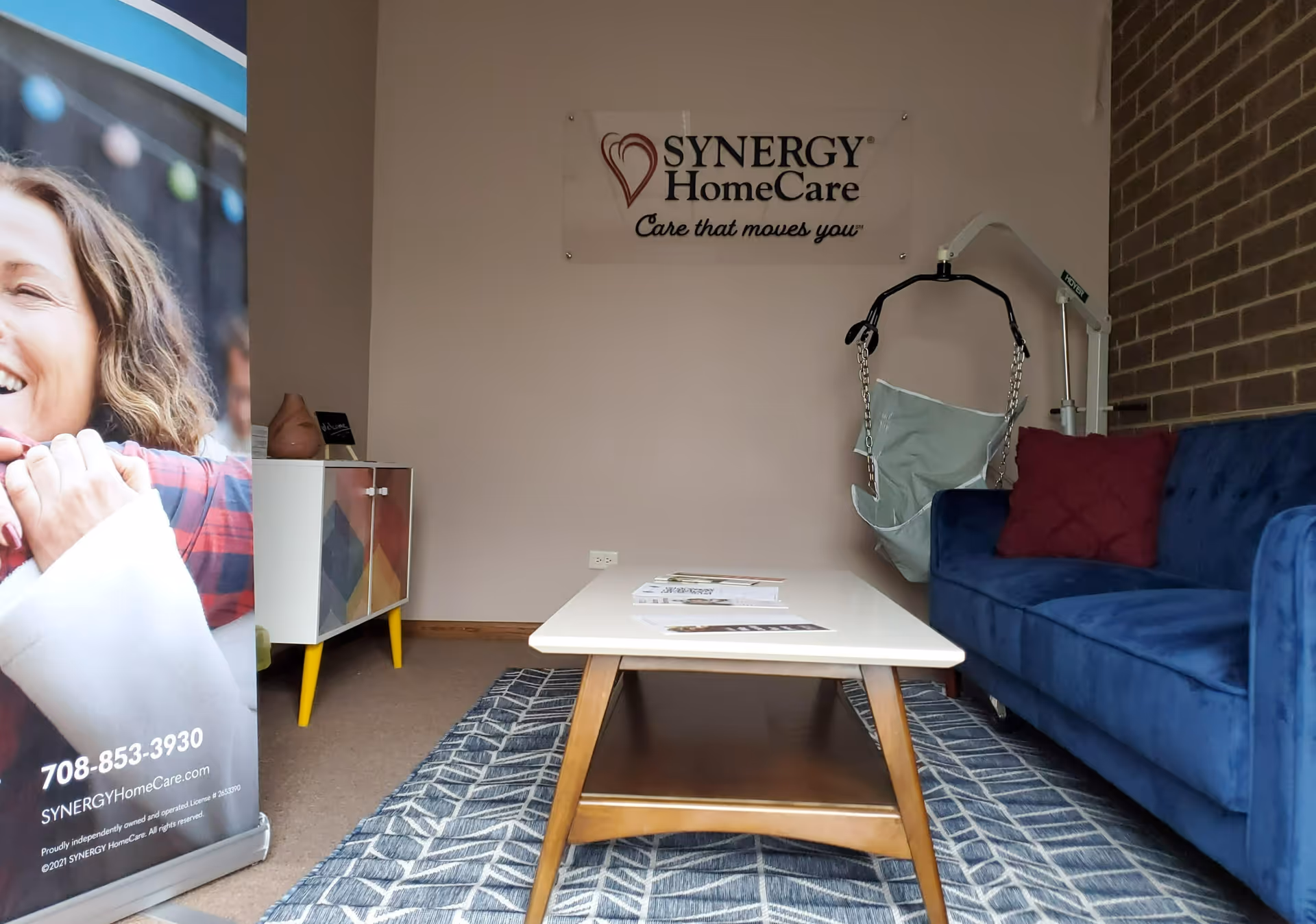An indoor waiting area with a blue sofa, coffee table, rug, and Synergy HomeCare banner.