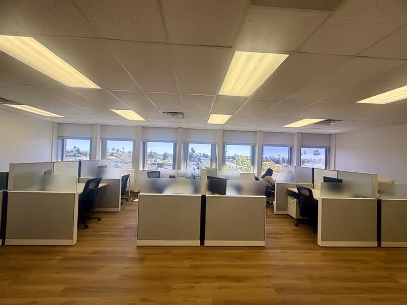 Open office area with cubicles, desks, and bright ceiling lights along back windows in facility