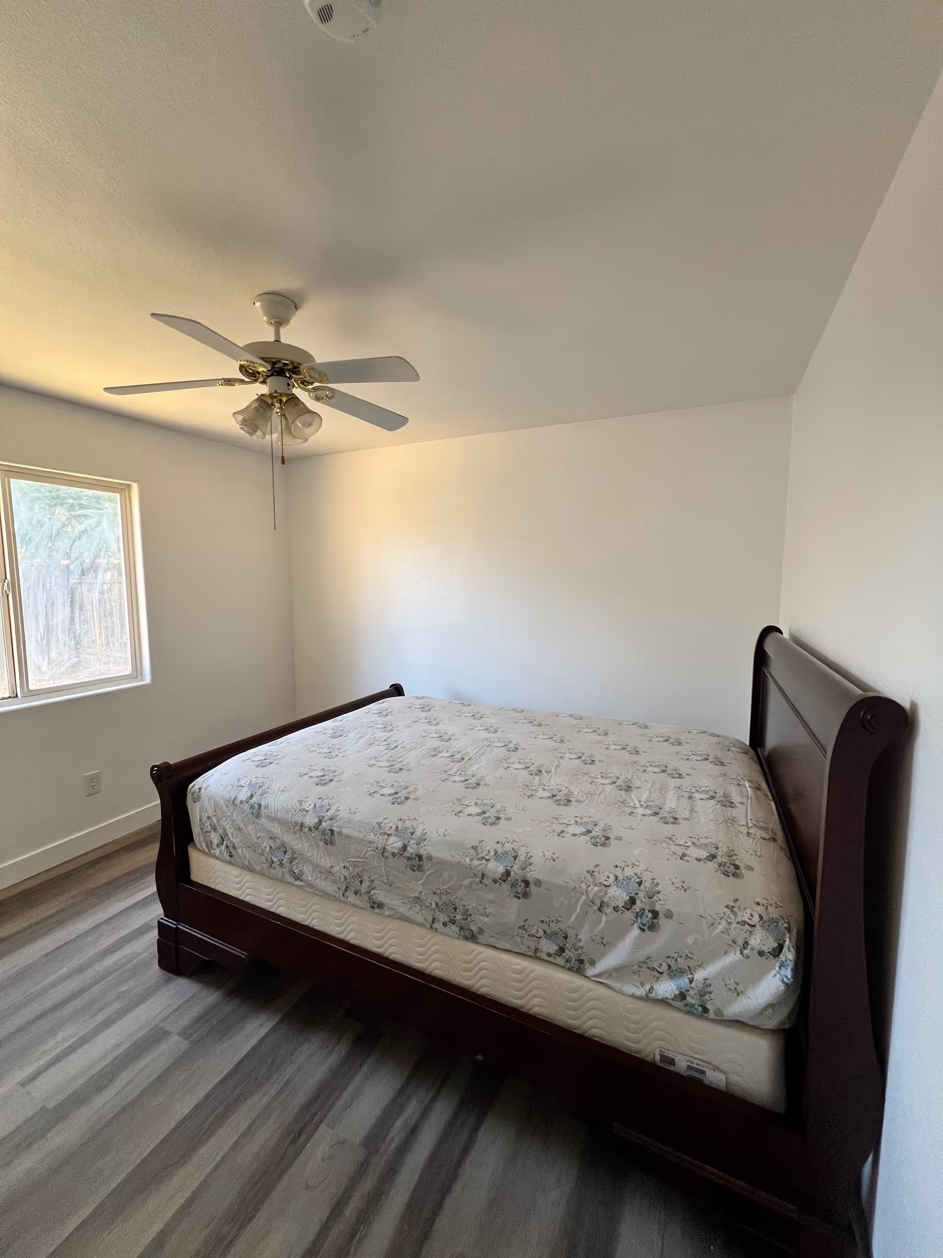 A simple bedroom with a dark wood bed, floral mattress, ceiling fan, and window.
