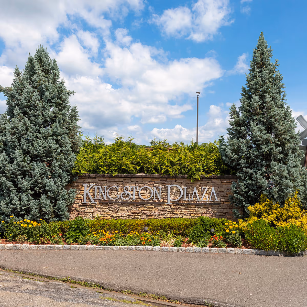 The Kingston Plaza sign sits among evergreen trees and blooming shrubs outside.