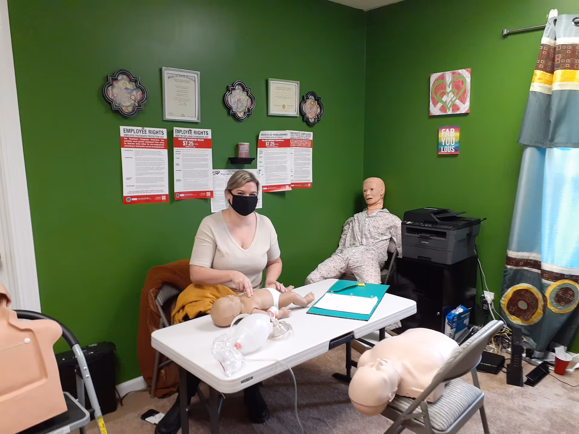 A training room with CPR mannequins, a seated instructor wearing a mask, and a green wall.