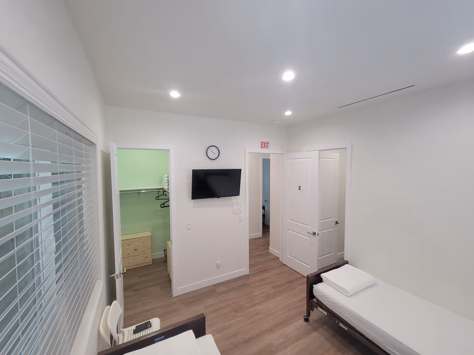 Senior facility bedroom with bed, blinds, wall-mounted TV, clock, and open closet.