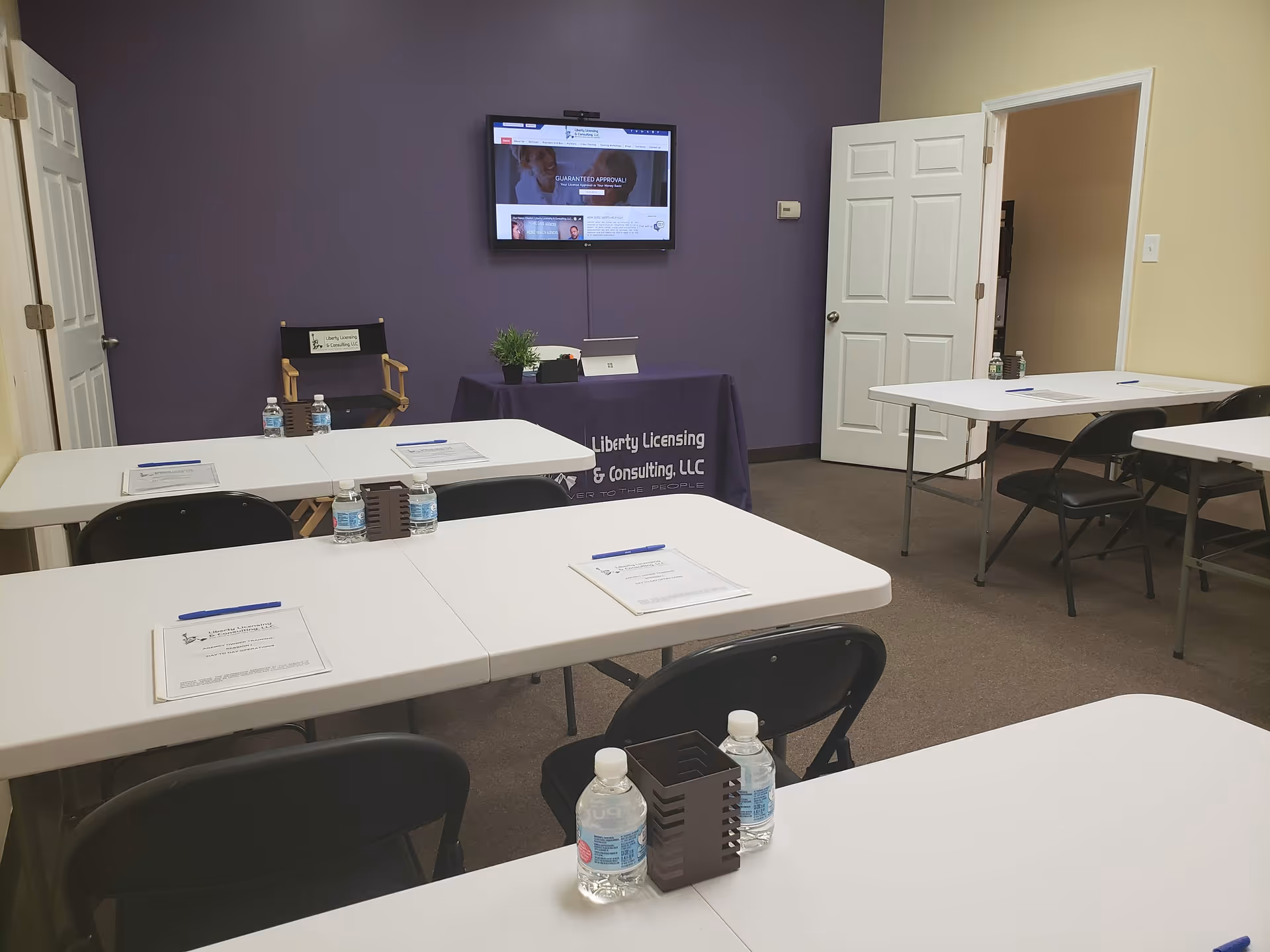 Conference room setup with white tables, chairs, water bottles, and a purple accent wall.