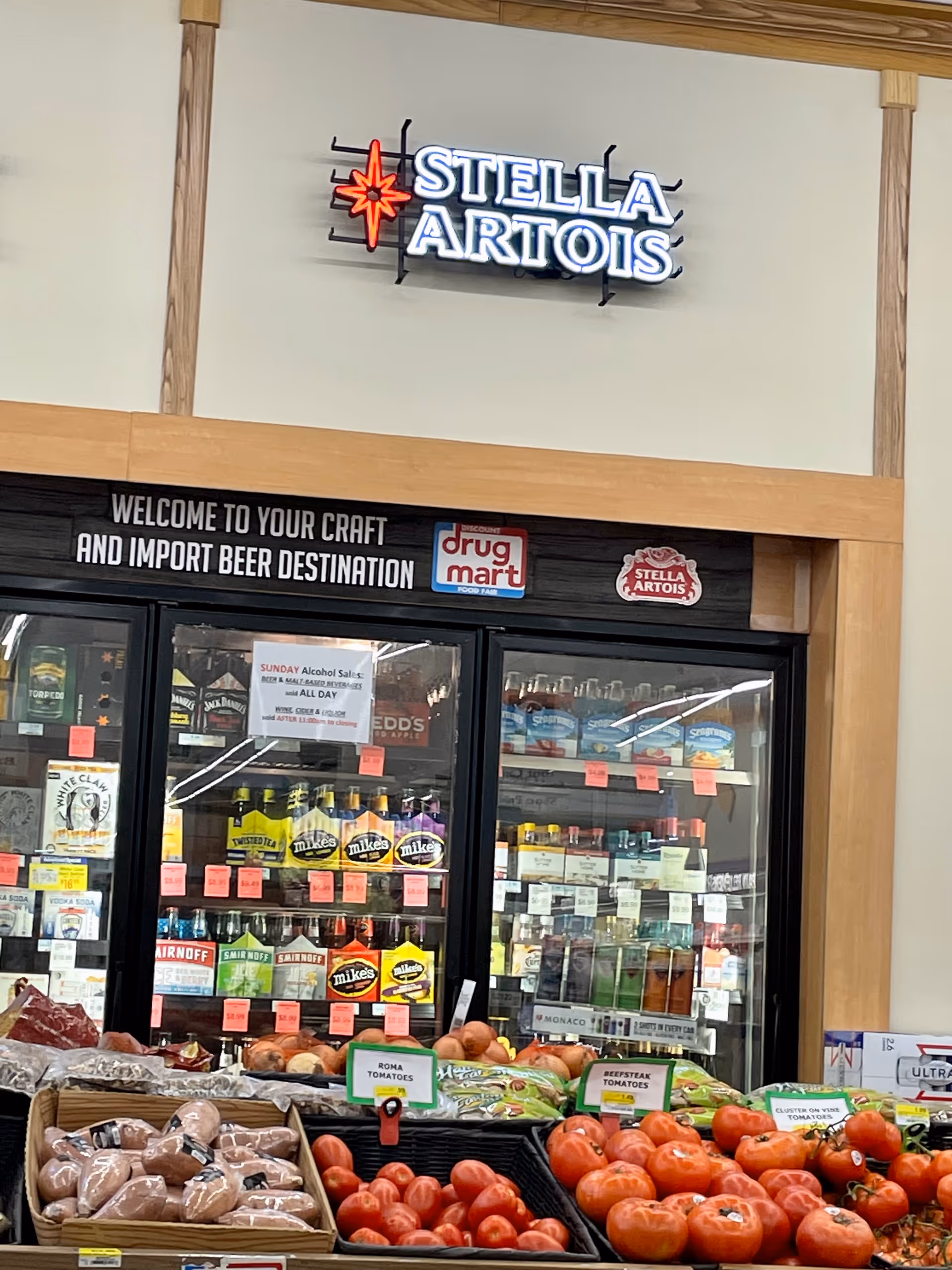 Neon Stella Artois sign above a beer display inside Discount Drug Mart grocery area.