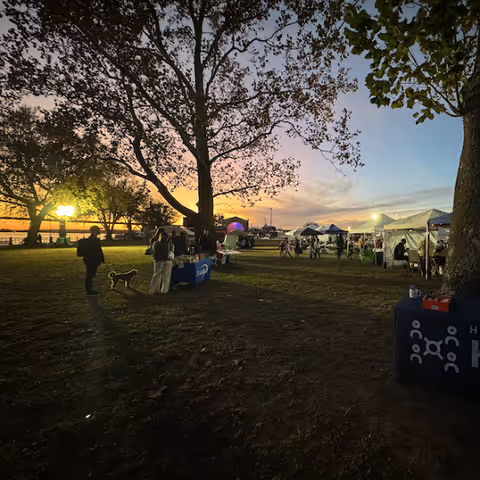 Sunset scene at an outdoor event with tents, trees, and people gathered on a lawn.