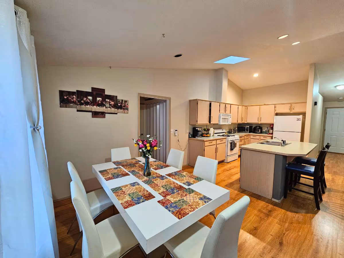 Bright dining and kitchen area with white table, six chairs, and flower centerpiece.