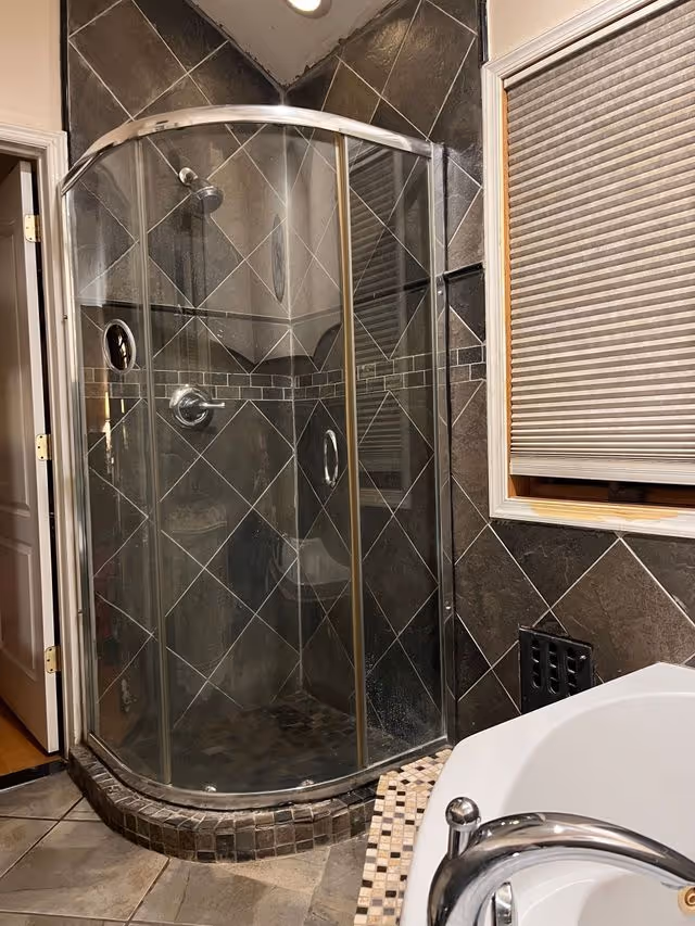 Curved glass shower enclosure with dark tiled walls beside a window in a bathroom.
